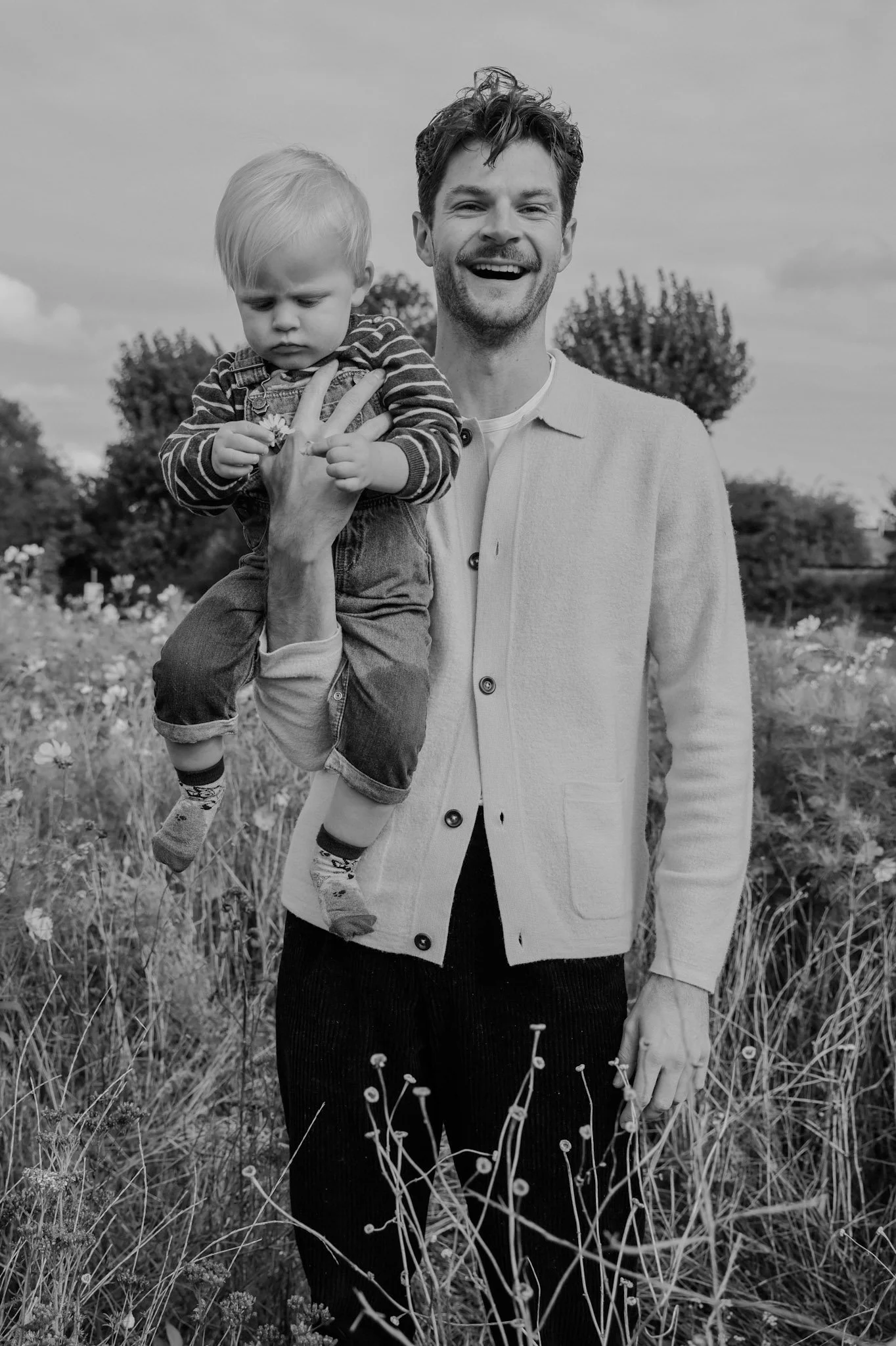 A smiling man holding a serious young boy in a field with flowers and trees in the background.