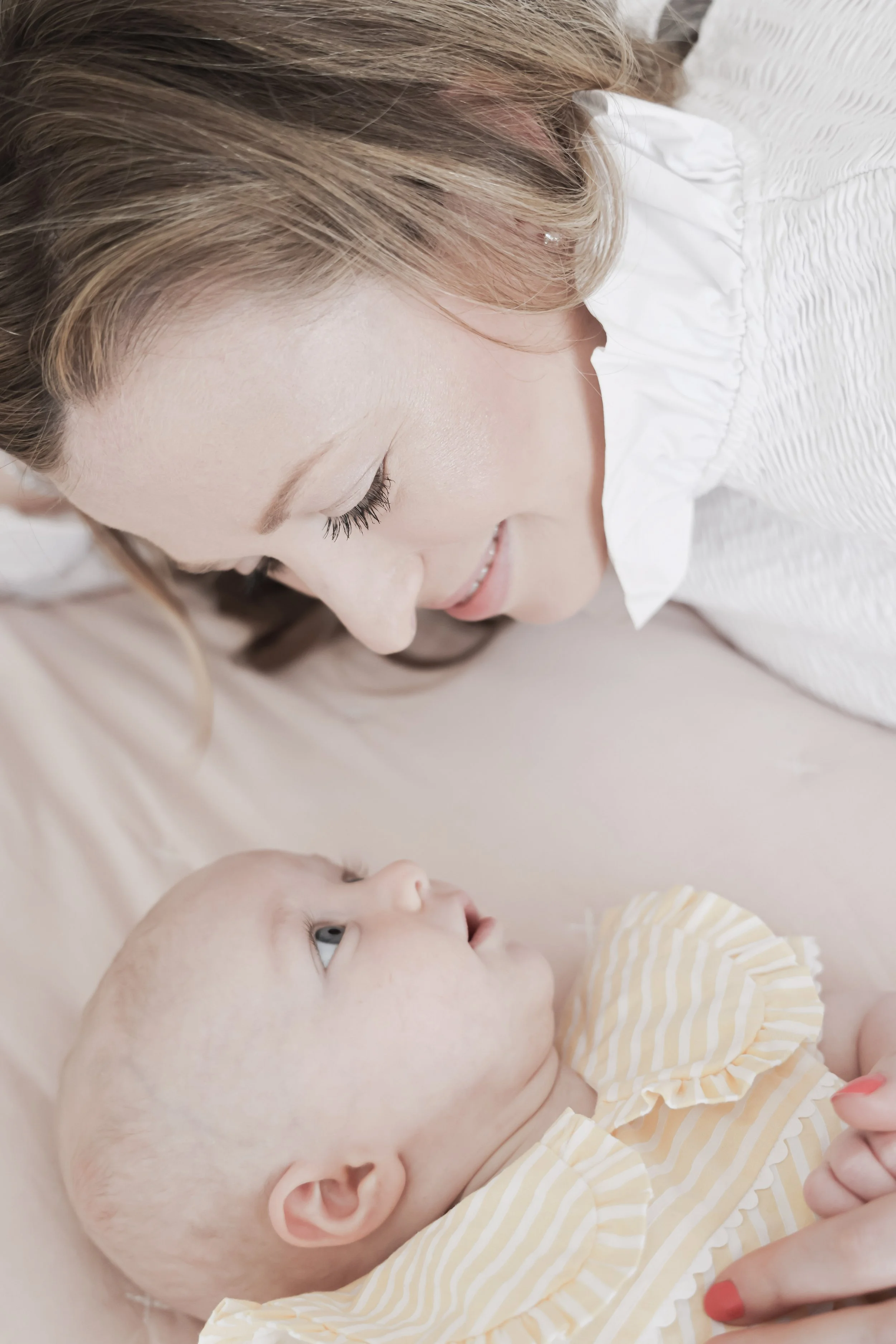 A woman smiles gently at a baby lying on a blanket, looking up at her.