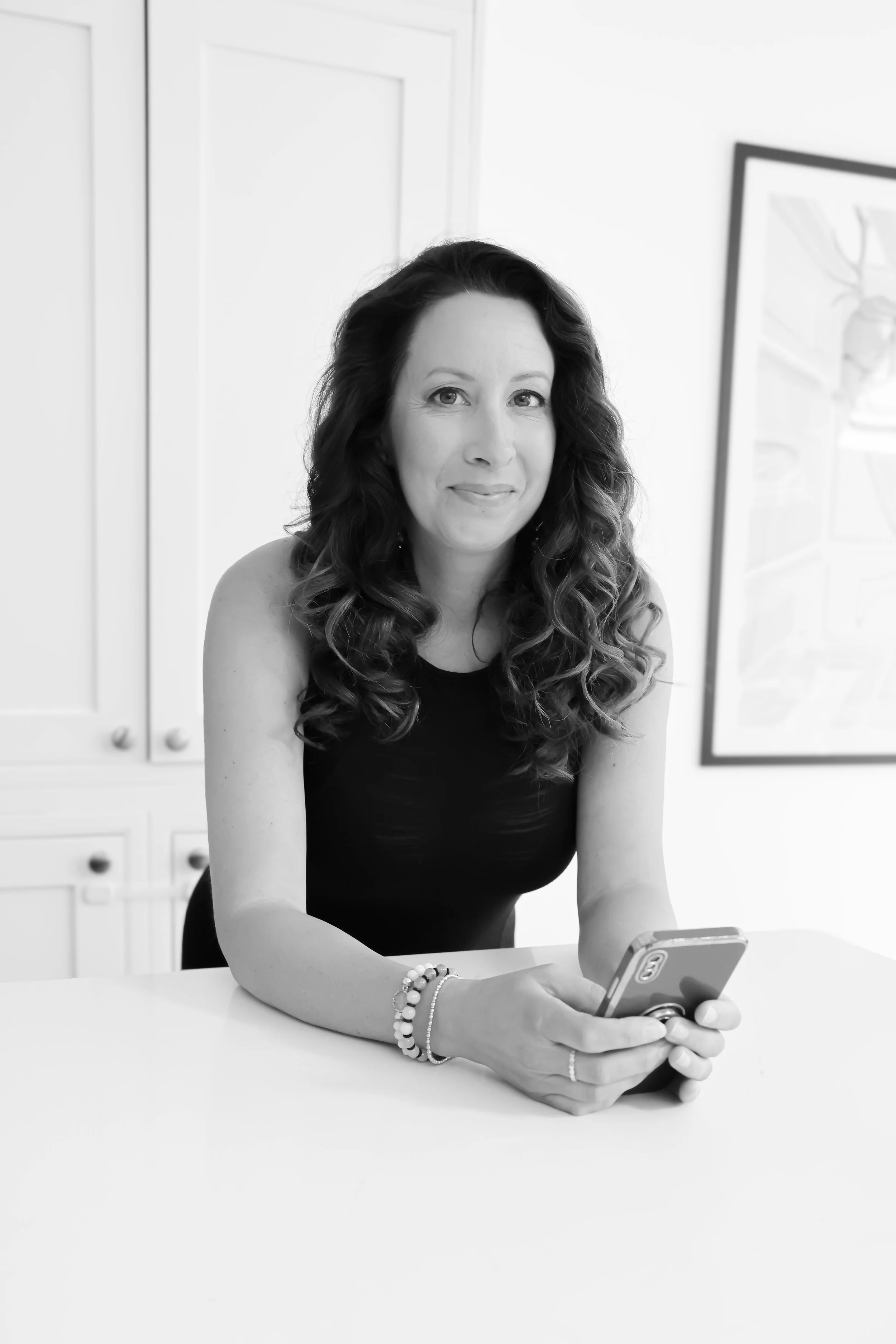 Black-and-white photo of a woman with curly hair sitting at a table, holding a smartphone, with kitchen cabinets and a framed picture in the background.