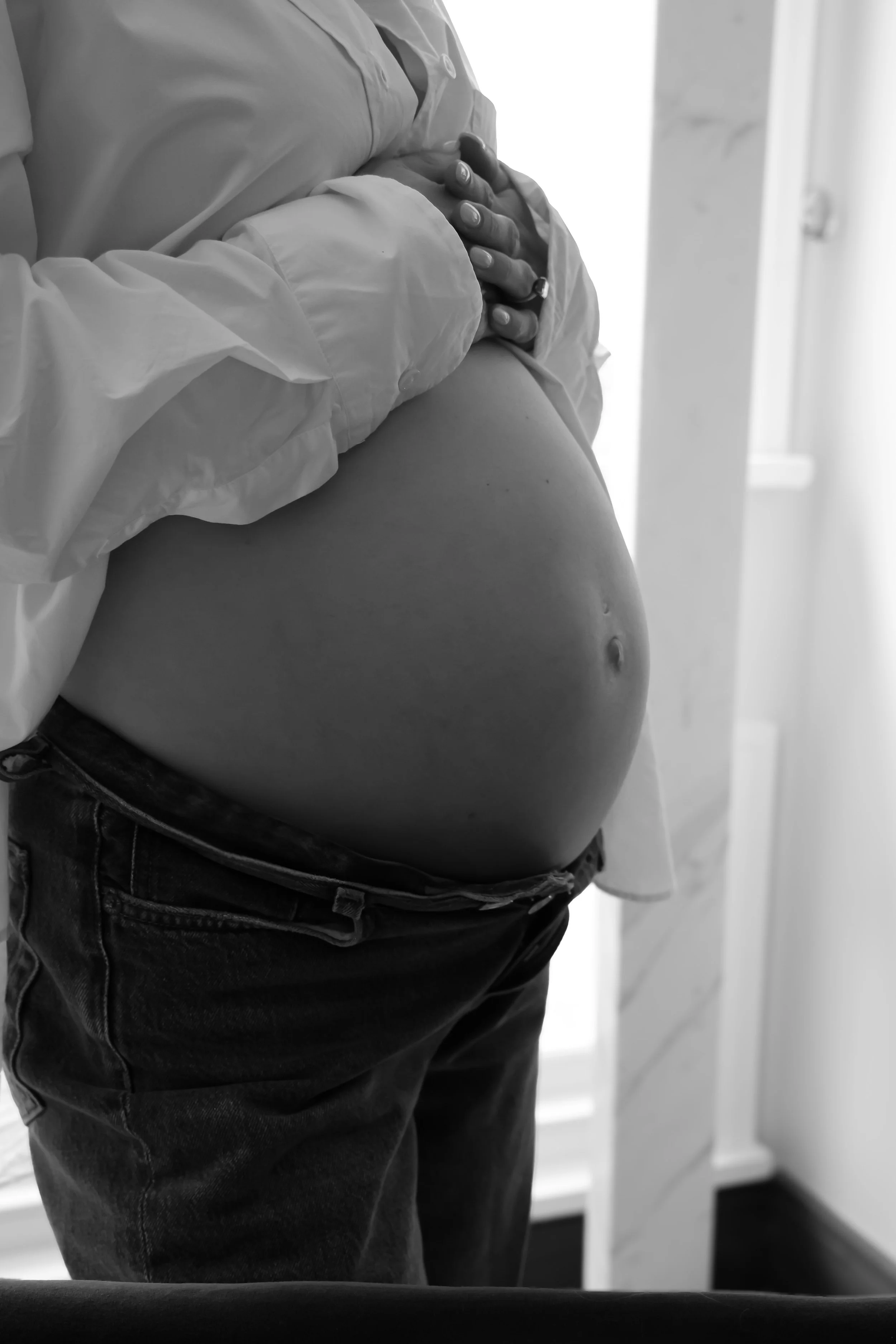 Black and white photo of a pregnant person holding their belly, standing indoors near a door.