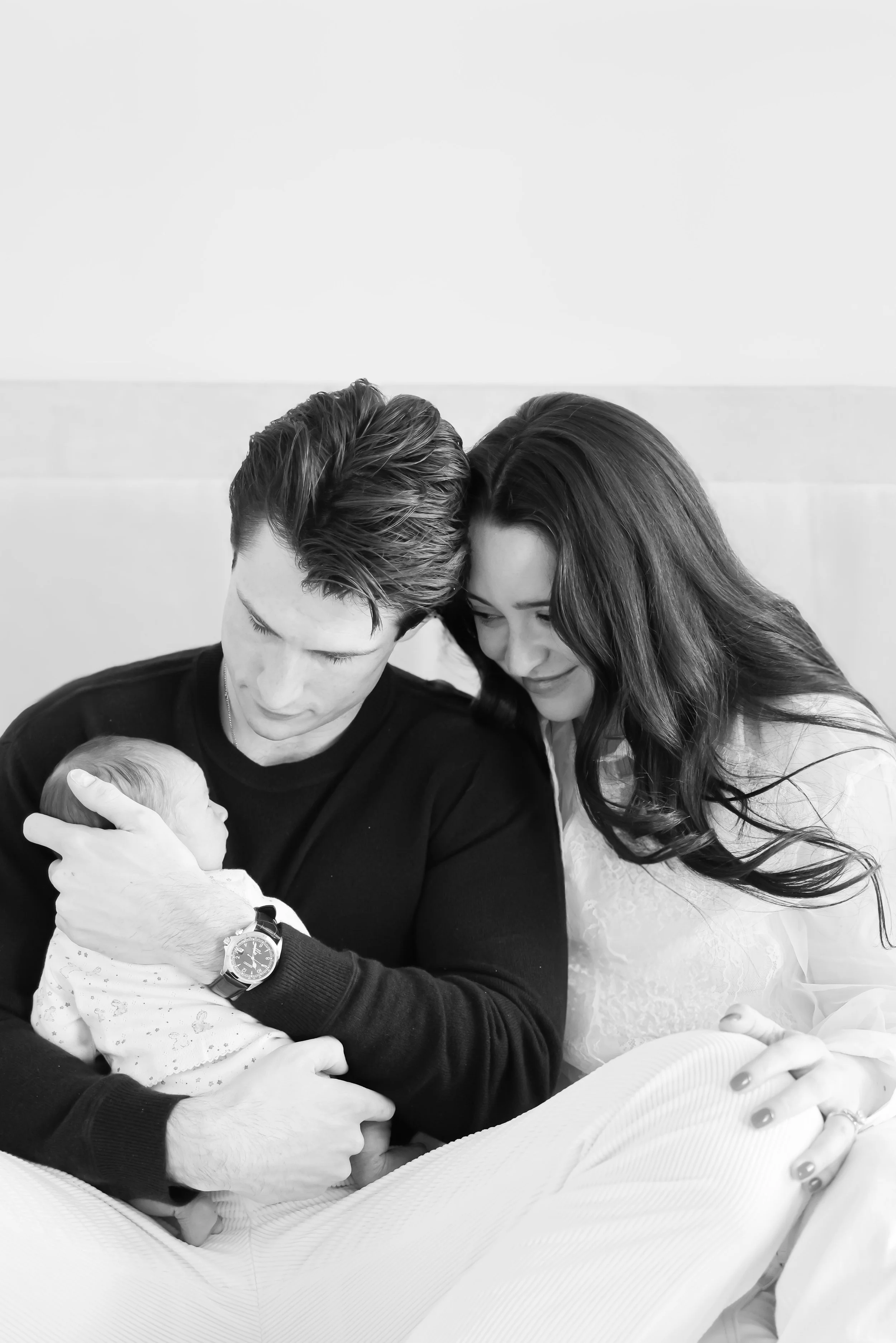 Black and white photo of a young man and woman looking at an infant. The man is holding the baby close to his chest while the woman leans in, both smiling softly at the child, who is swaddled in a blanket.