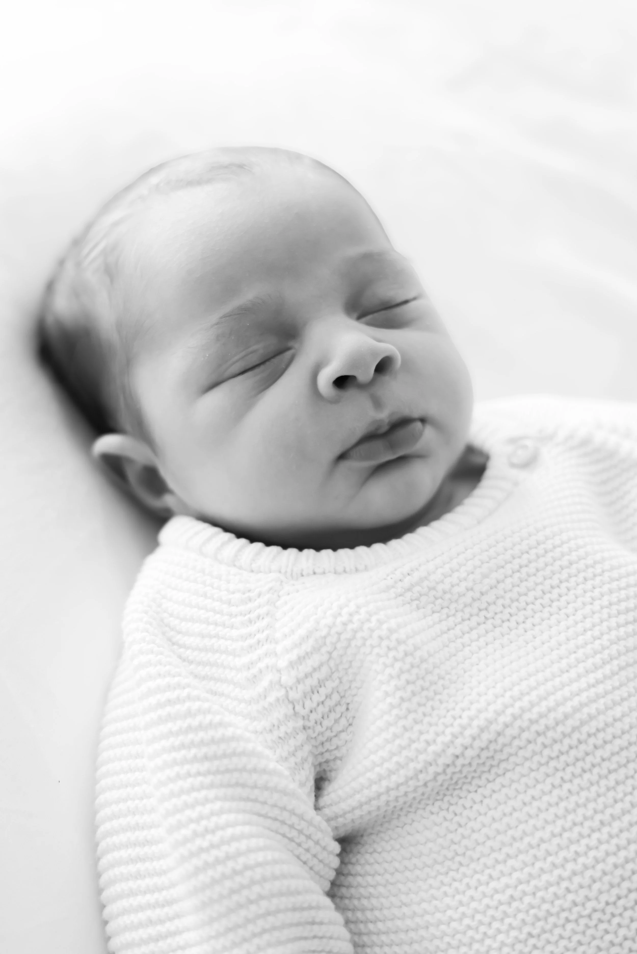 Black and white photo of a sleeping infant with closed eyes, lying on a soft surface, wearing a knitted sweater.