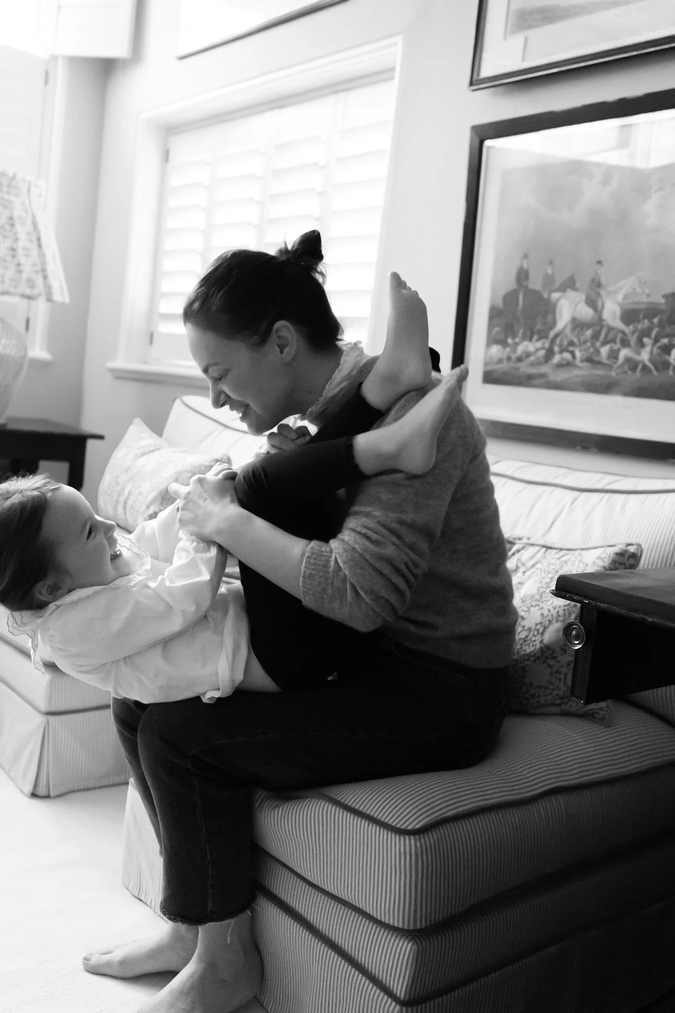 A woman and a young girl playing on a couch in a living room, smiling and laughing together, black and white photo.