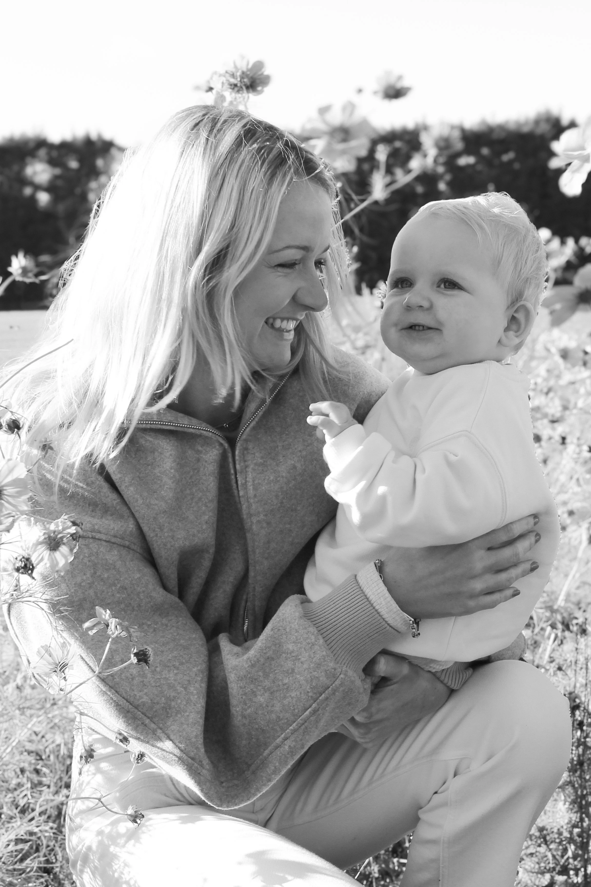 A woman and a young child smiling and enjoying a moment outdoors among blooming flowers, captured in black and white.
