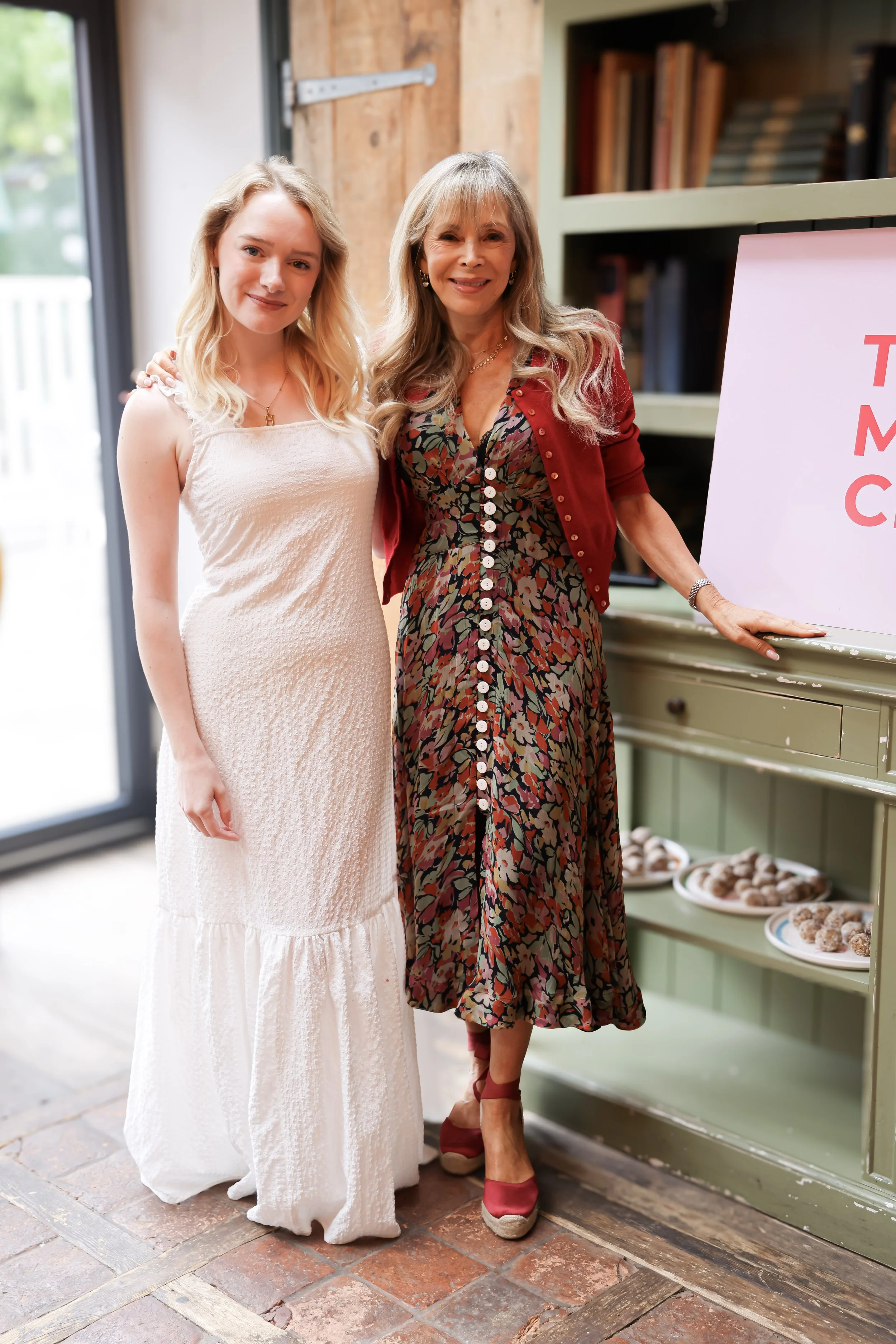 Two women standing inside a room. The woman on the left wears a long, white, textured dress, and the woman on the right wears a floral dress with a red cardigan. They are standing next to a green cabinet with plates of food and a pink sign. The setting appears to be a casual gathering or event.