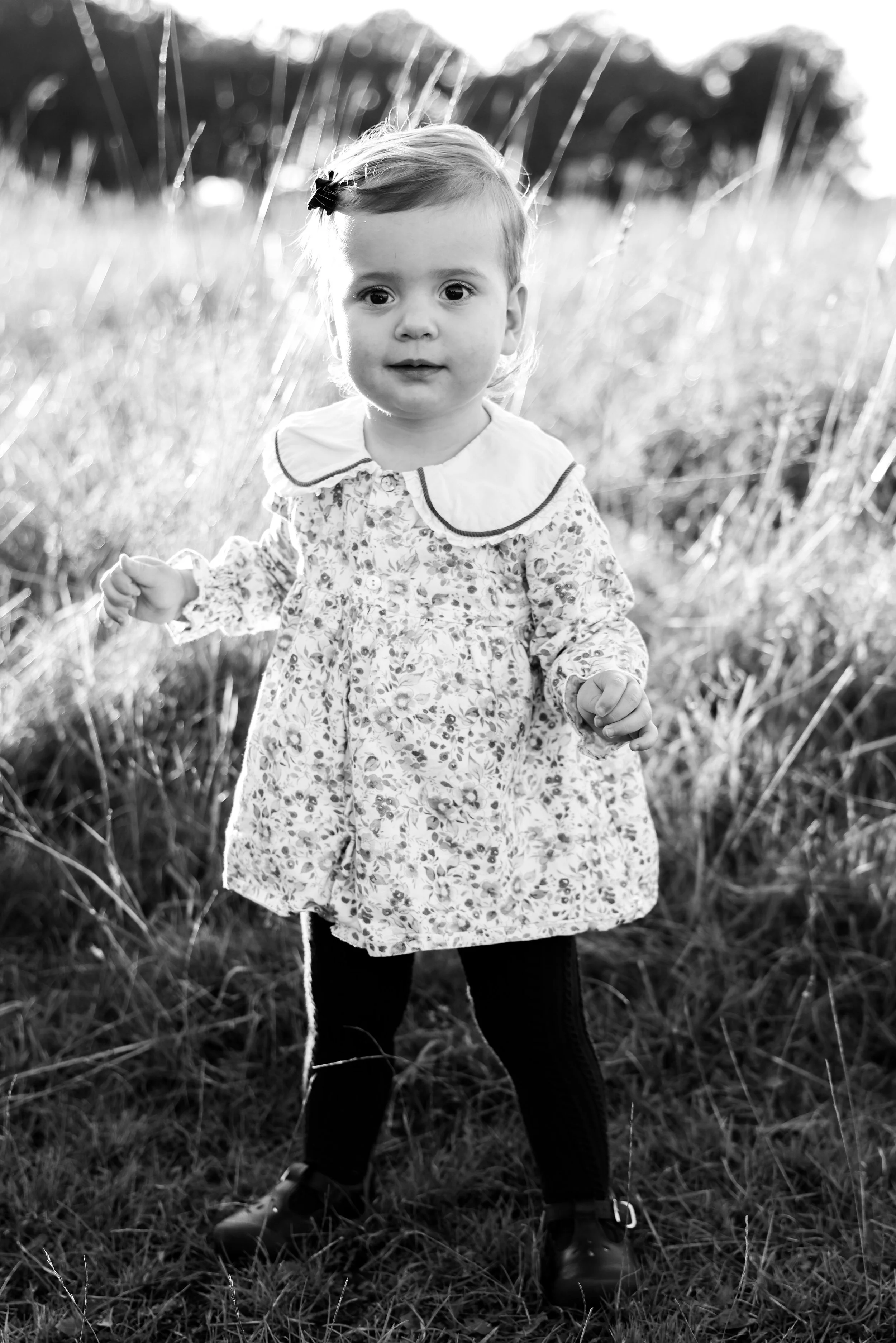 A young girl standing outdoors in a grassy field, wearing a floral dress, dark leggings, and shoes, with her hair held back by a small bow, captured in black and white.