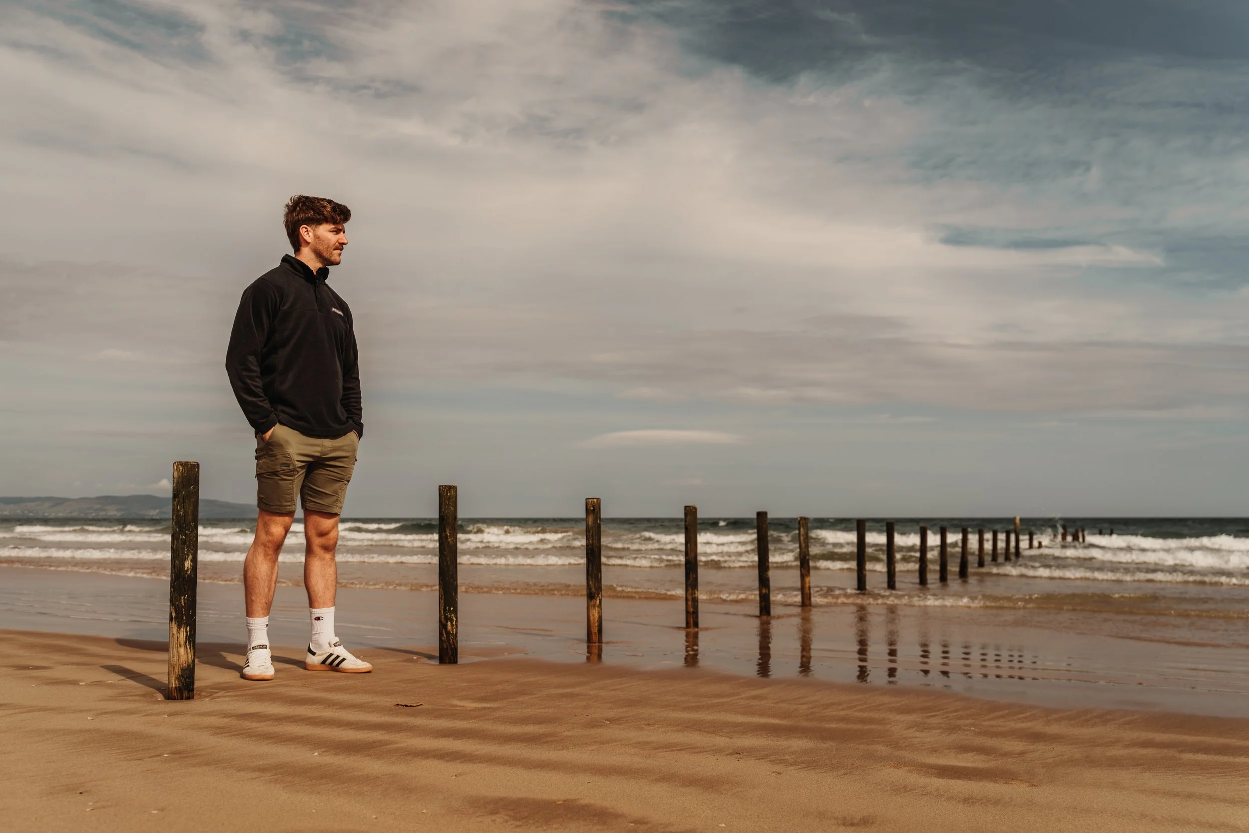 A man standing on a sandy beach near wooden posts, looking towards the ocean under a cloudy sky.