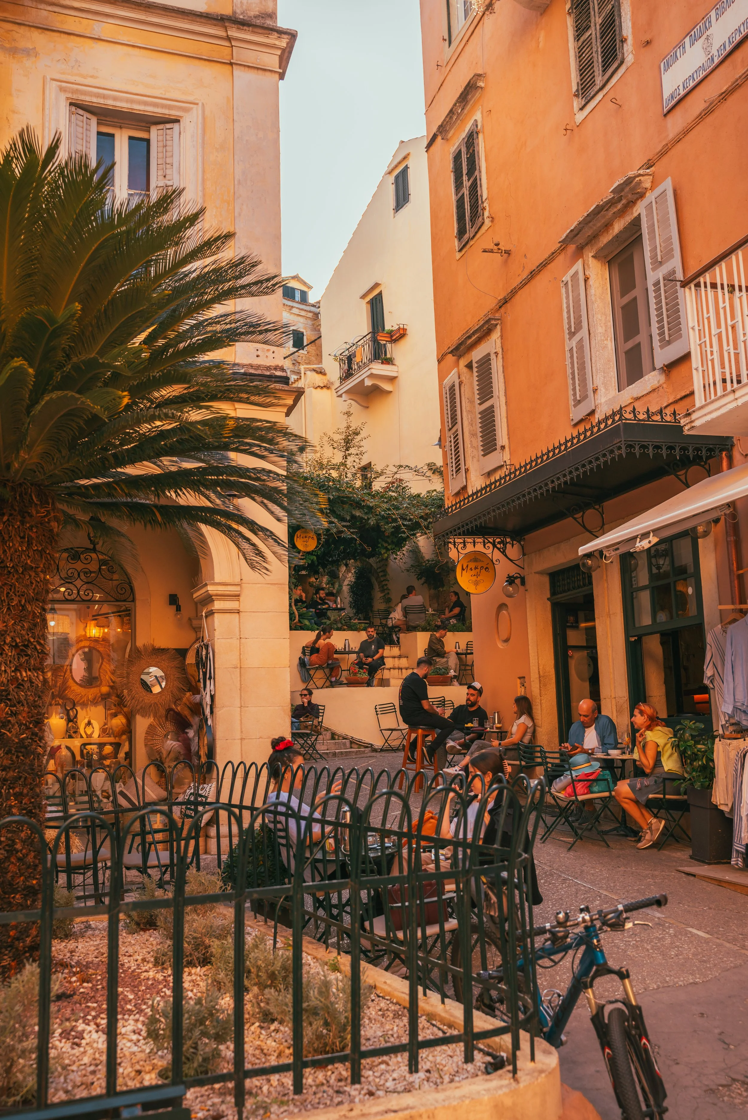 A lively outdoor café scene in a European alley with people sitting at tables, stairs leading up with more patrons, colorful buildings with shutters, a palm tree, and bicycles parked in front.