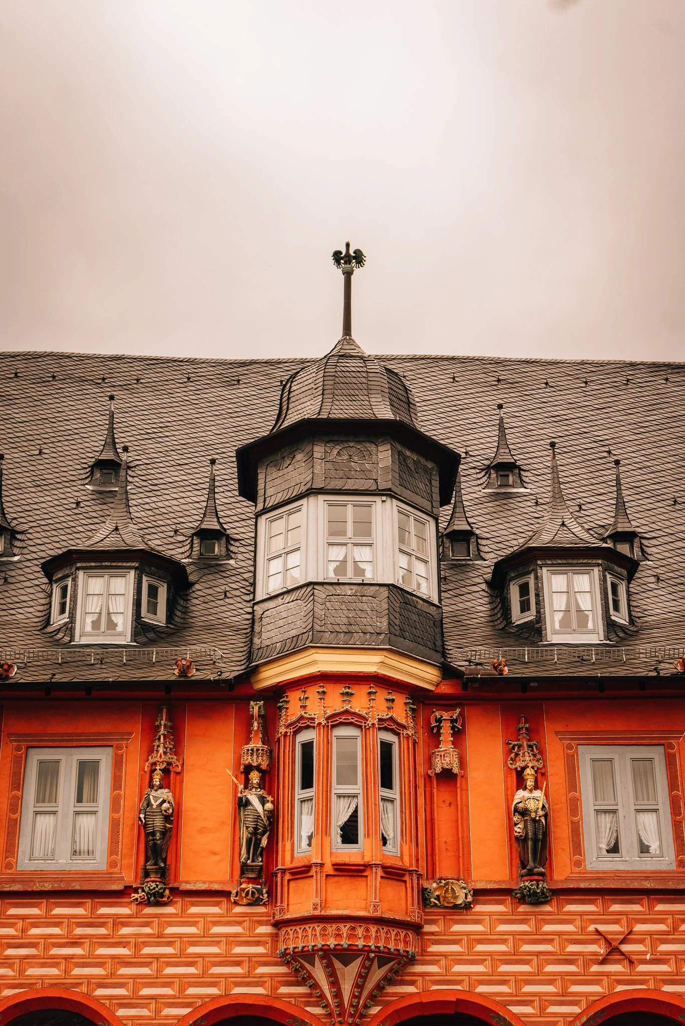 The decorative upper facade of a historic building featuring ornate statues, intricate window designs, and a steep, shingled roof with multiple small dormer windows and a central tower topped with a weather vane.