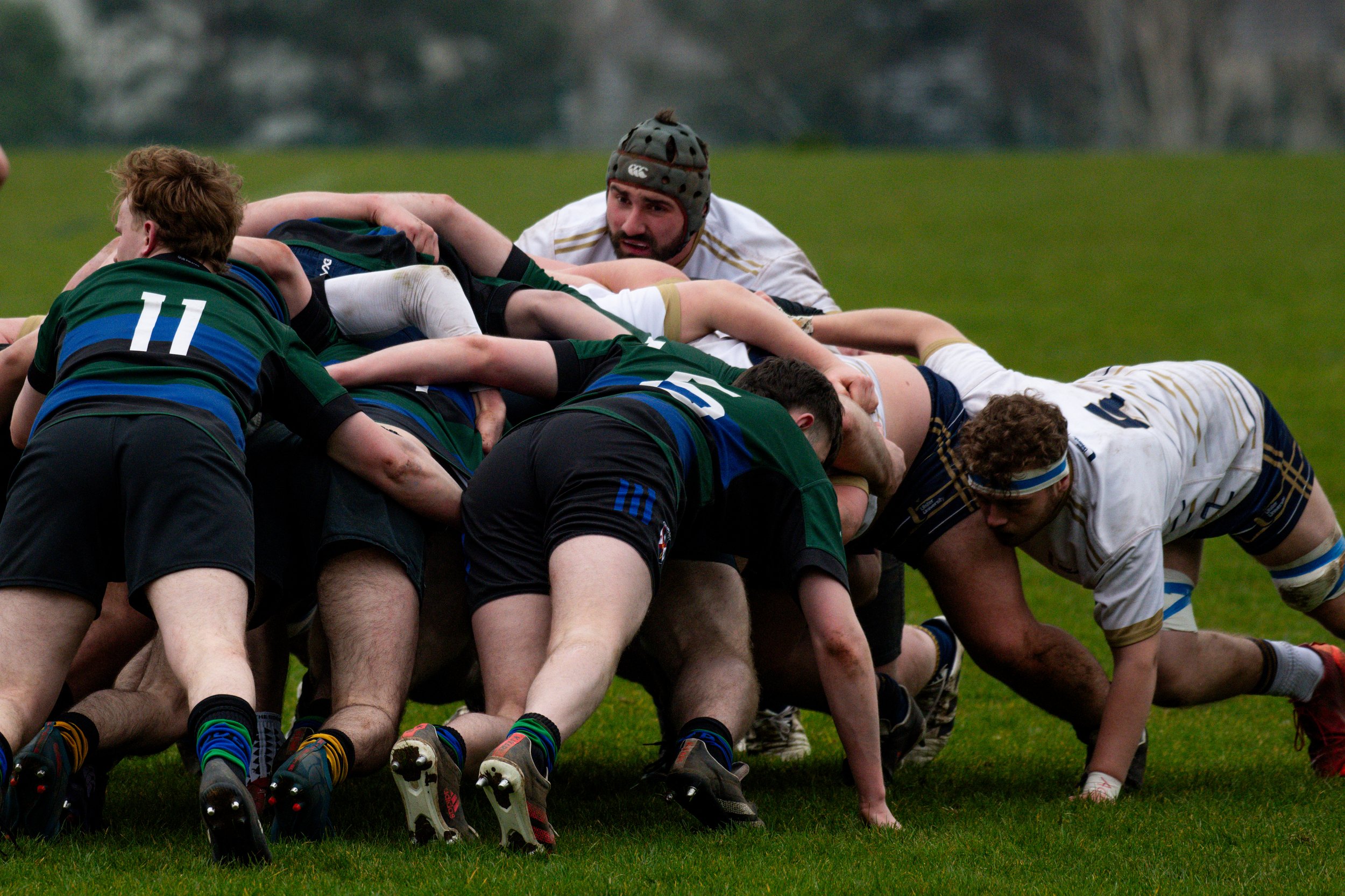 Rugby players in a scrum on a grassy field, with one player wearing a helmet and others in uniforms with numbers.