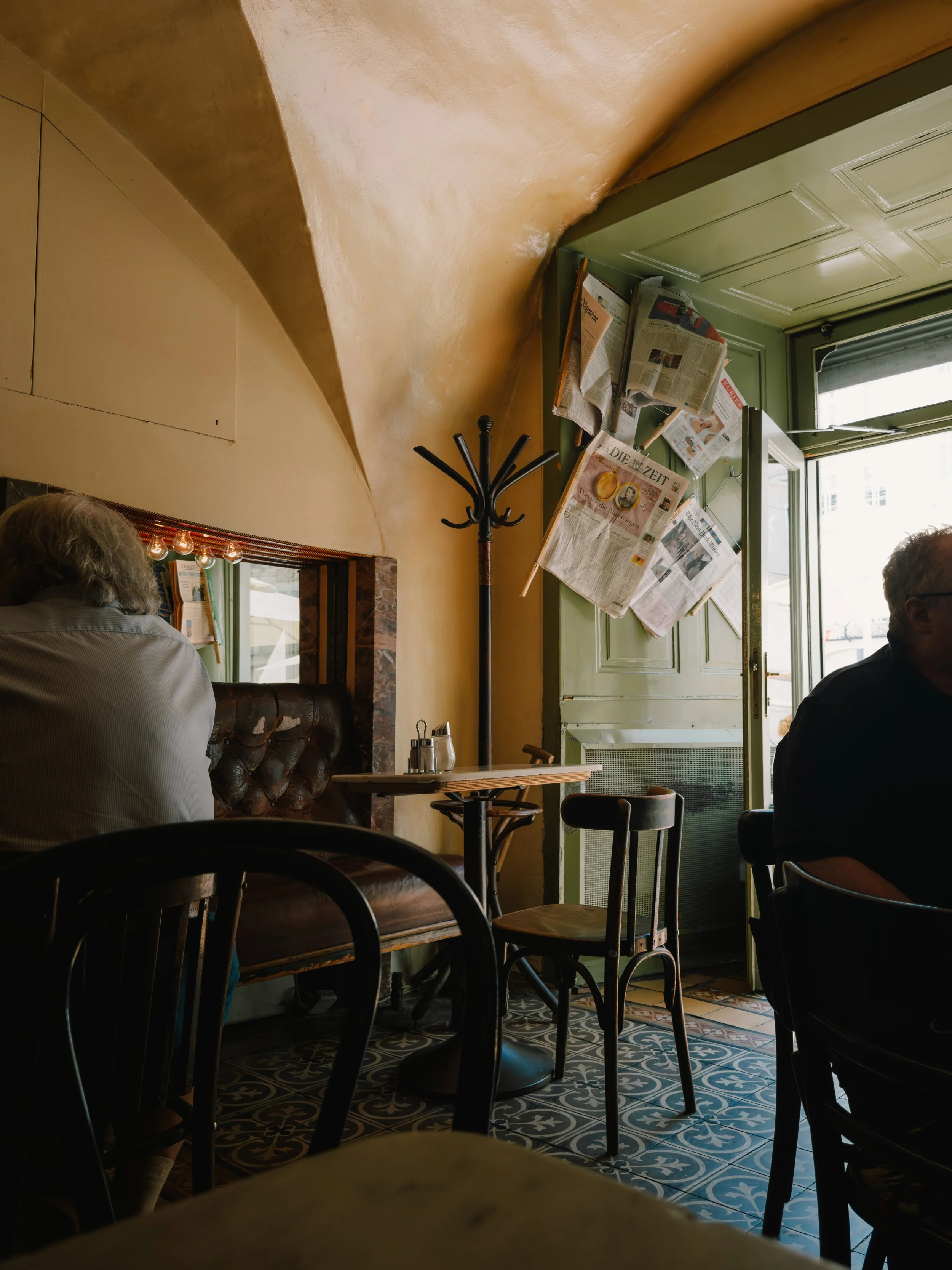 Interior of a cozy café with a small table and wooden chairs, newspapers pinned to a door, soft lighting, and patterned tile floors.