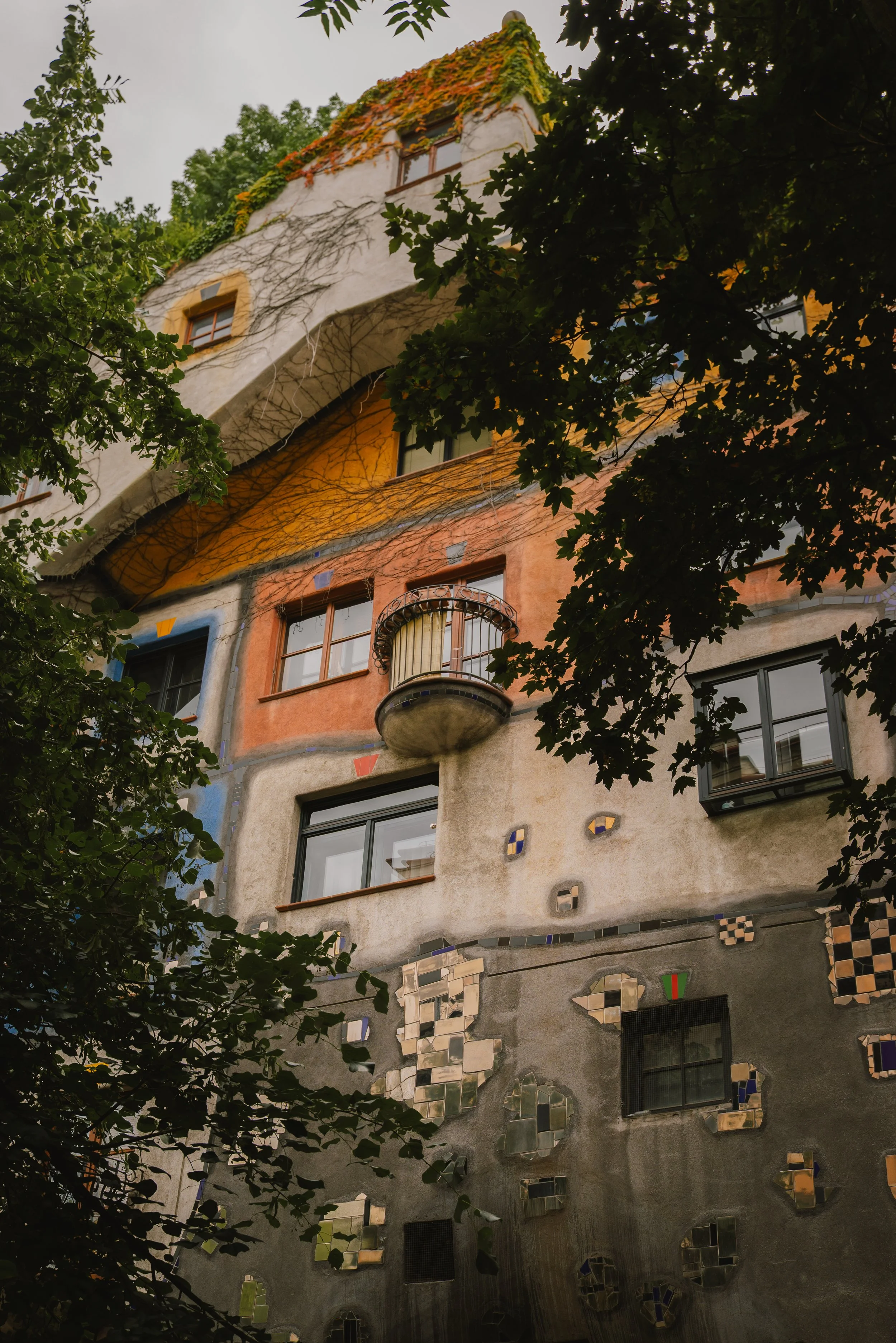 Colorful, architecturally unique building with irregularly shaped windows, balconies, vivid colors, and decorative mosaic details, framed by leafy trees.