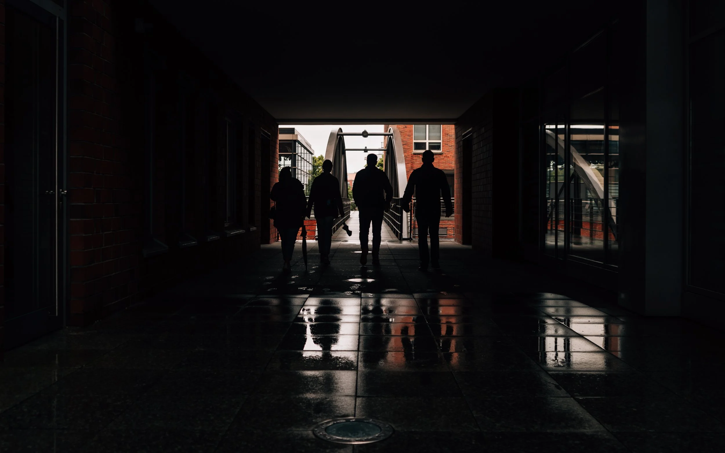 Four silhouettes of people walking in an urban passageway towards an outdoor area with a bridge or stairs, with reflections on the wet floor.