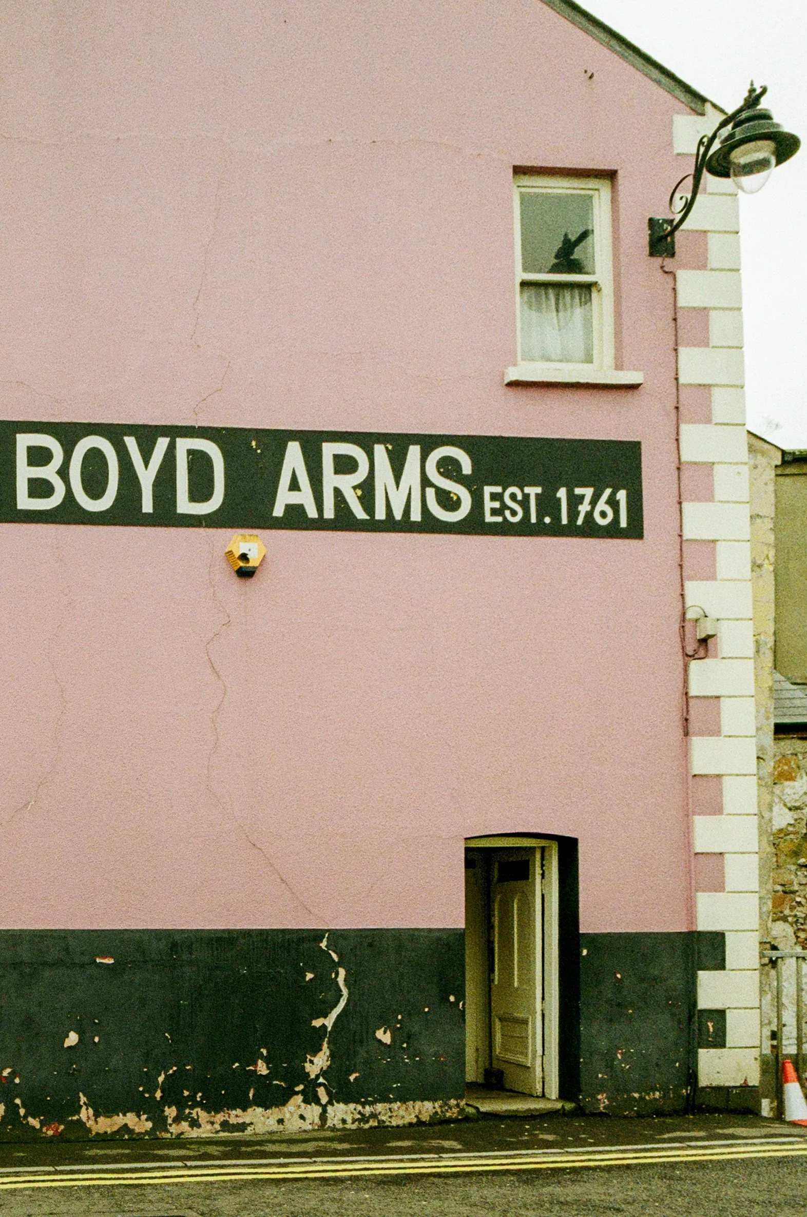 A pink building with a sign that says Boyd Arms, established in 1761, with a small window and an open door at ground level, a decorative wall light, and a small corner window on the upper floor.