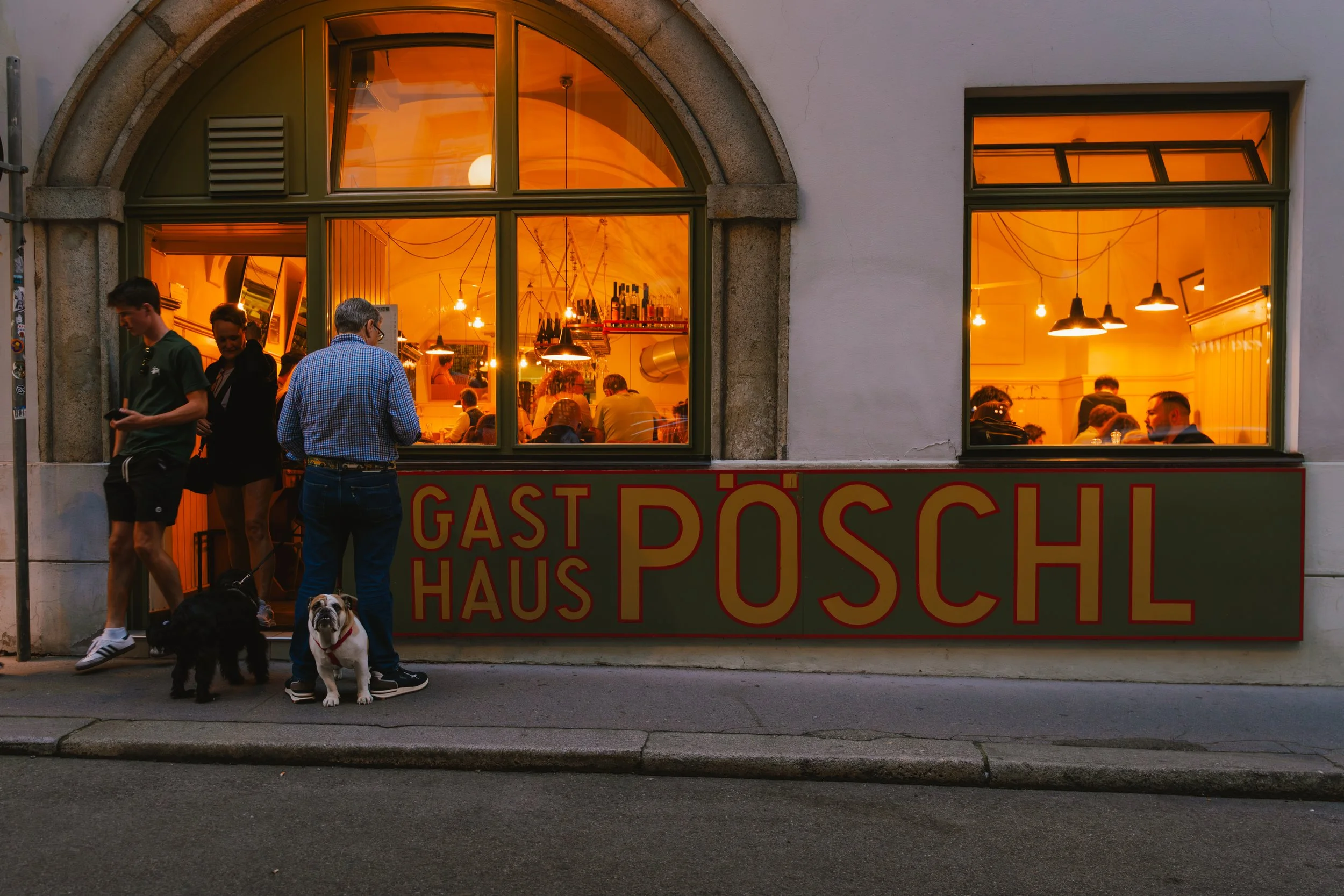 People outside a restaurant named Gast Haus Pöschl looking inside through large windows, with some dogs on the sidewalk.