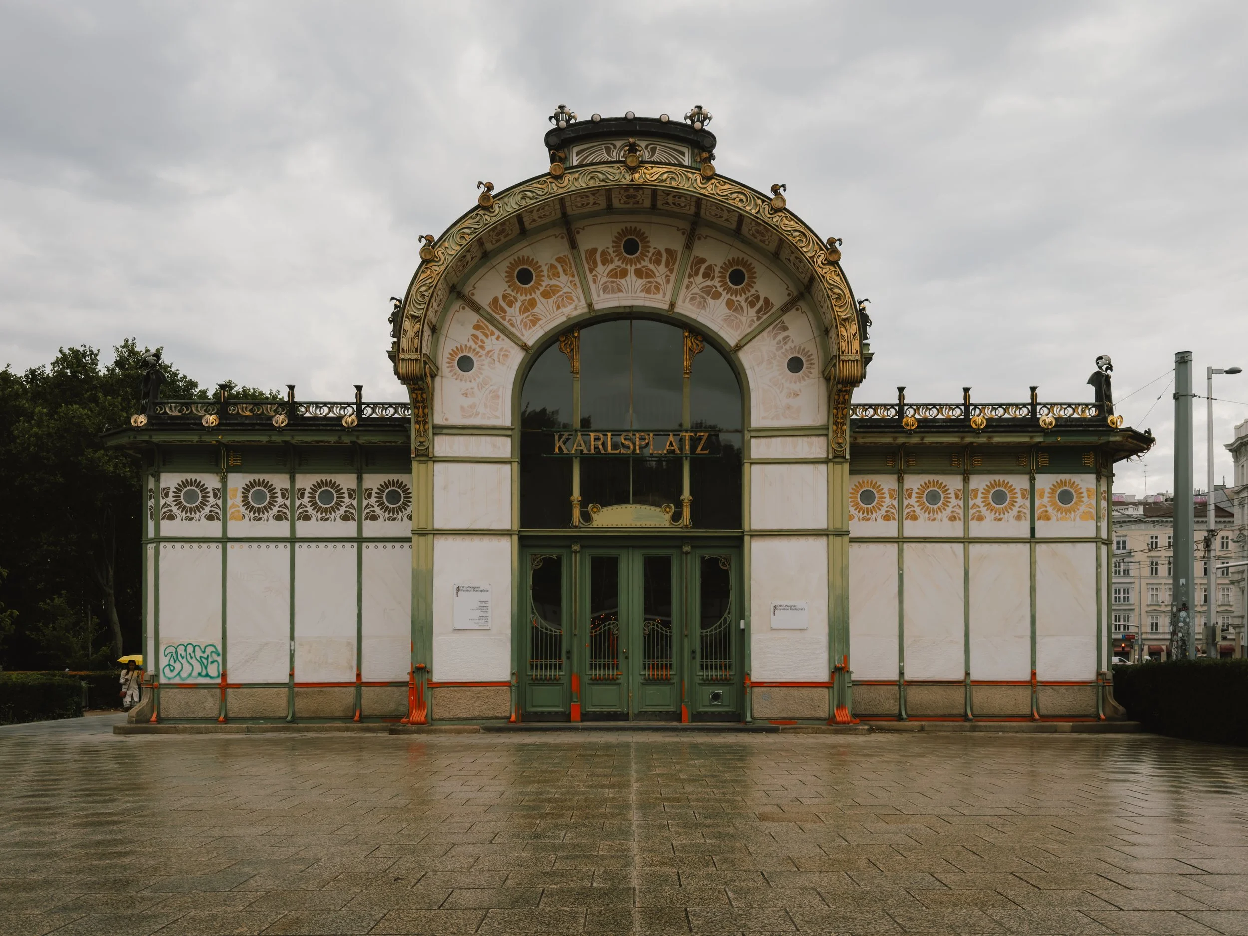 Historic ornate building with a sign reading 'Karlsplatz' in a European city, cloudy sky, and wet pavement.