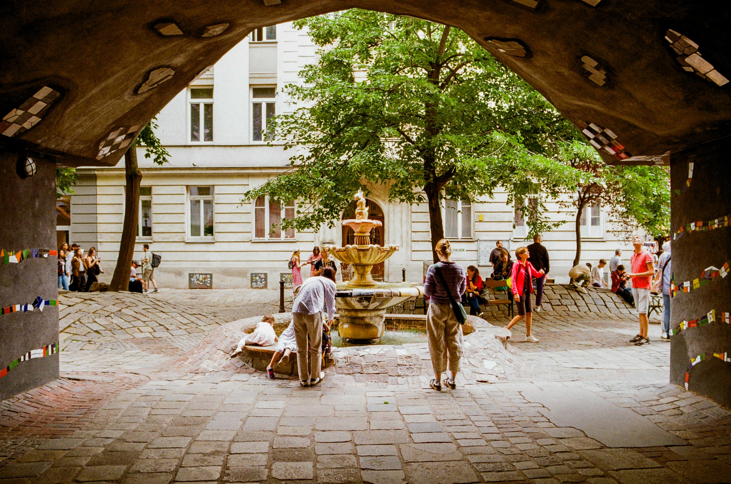 View from under a bridge or archway of a city square with people gathered around a central fountain, large trees, and a white building in the background.