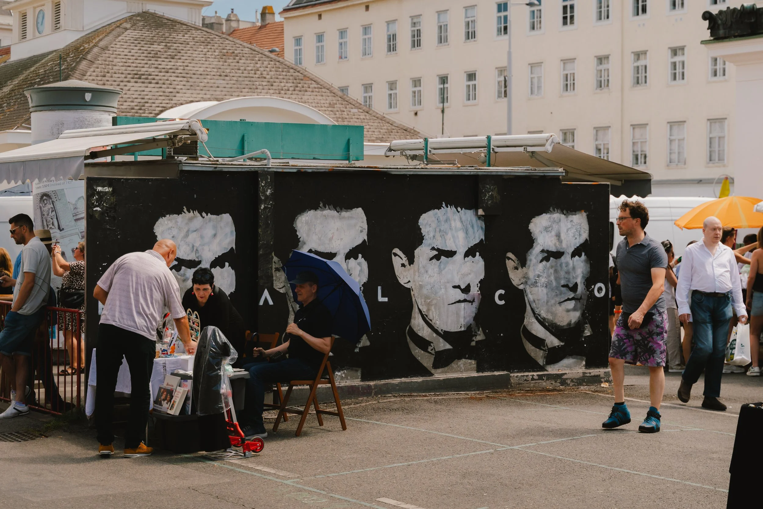 A mural of four men's faces on a black wall at an outdoor market, with people browsing and walking by, some under umbrellas, in an urban setting.