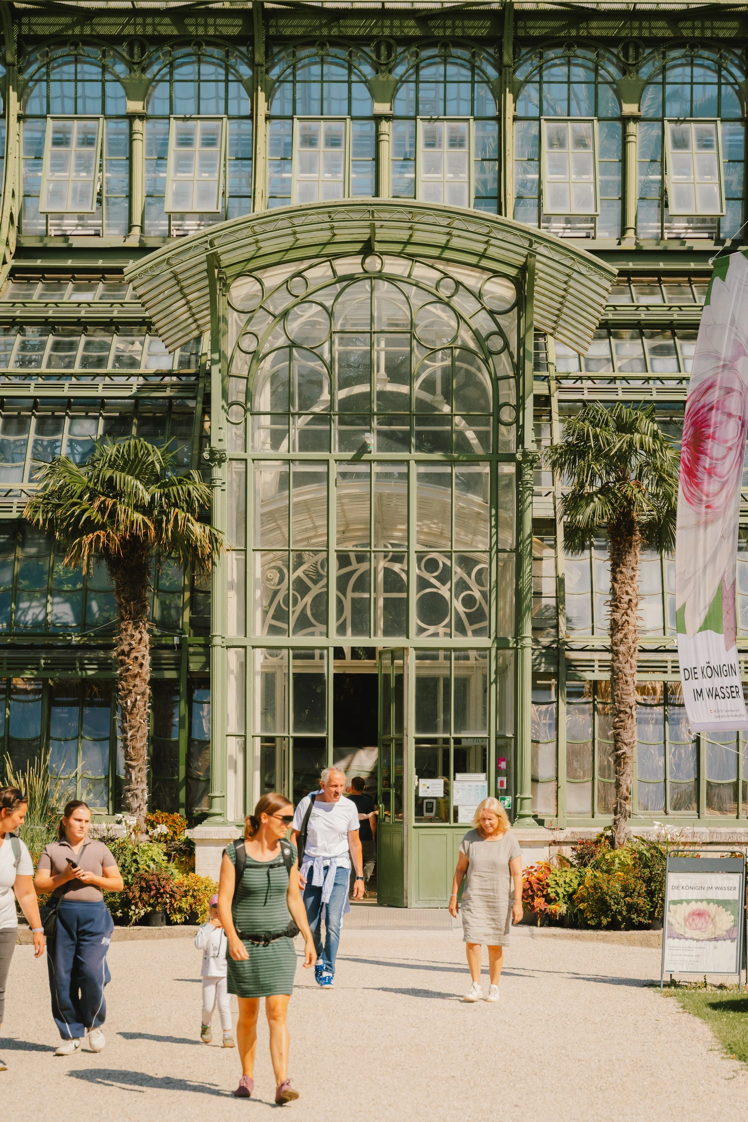 Visitors walking in front of a large glass greenhouse with palm trees and colorful plants outside.
