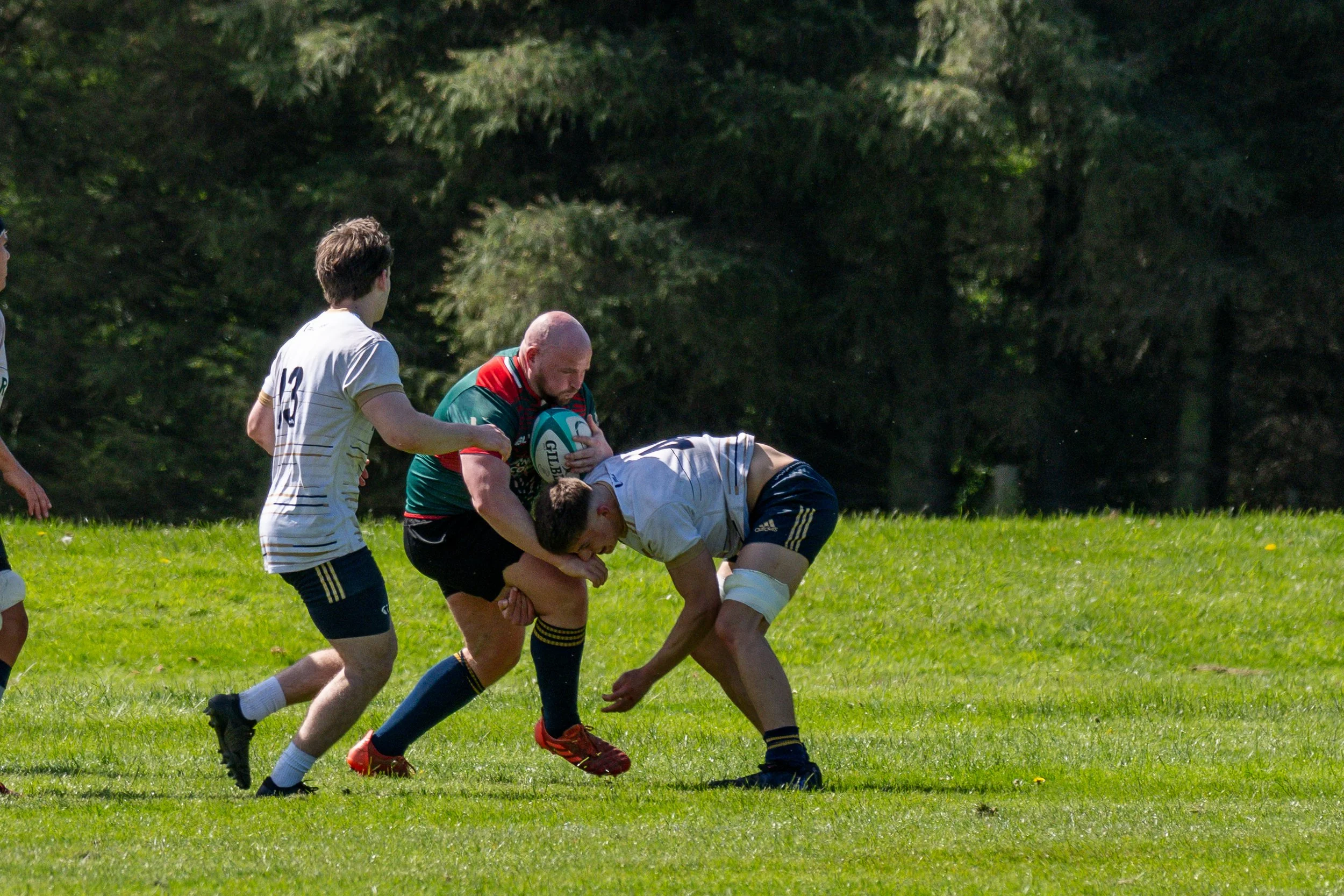 Rugby players contesting for the ball on a grassy field, with one player holding the ball and other players trying to tackle him, against a background of trees.