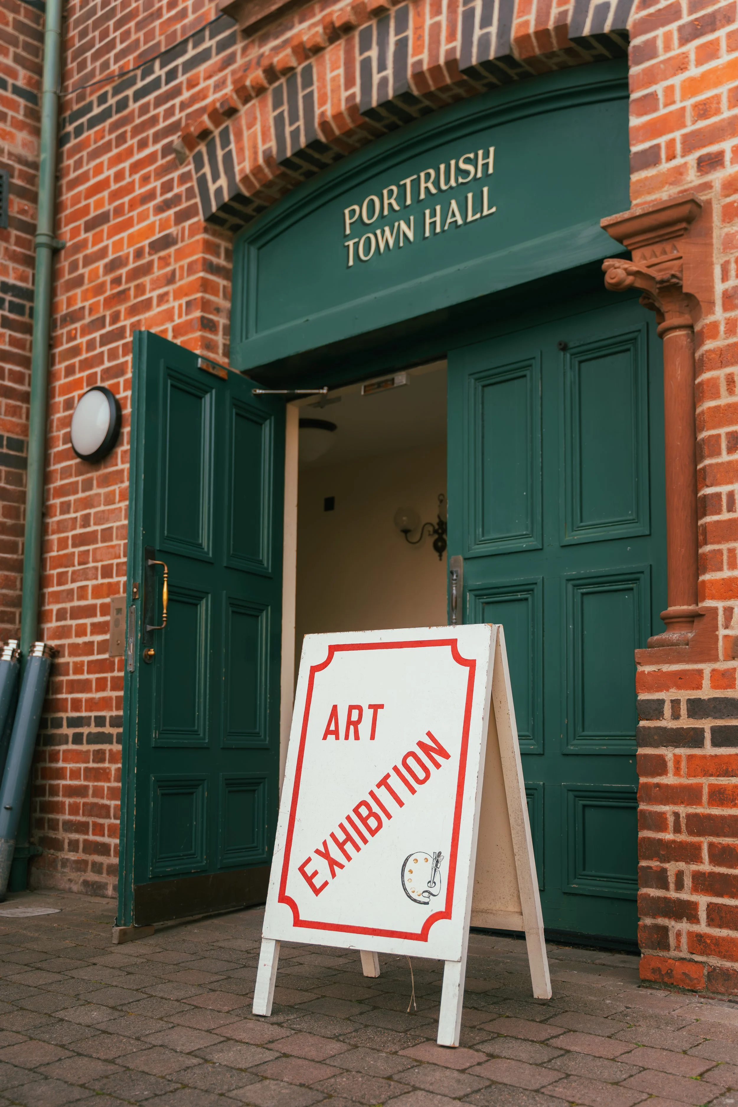 Green double doors of Portrush Town Hall with a white sign outside advertising an art exhibition, featuring a red border, the words 'ART EXHIBITION', and a small artist's palette illustration, set against a brick building.