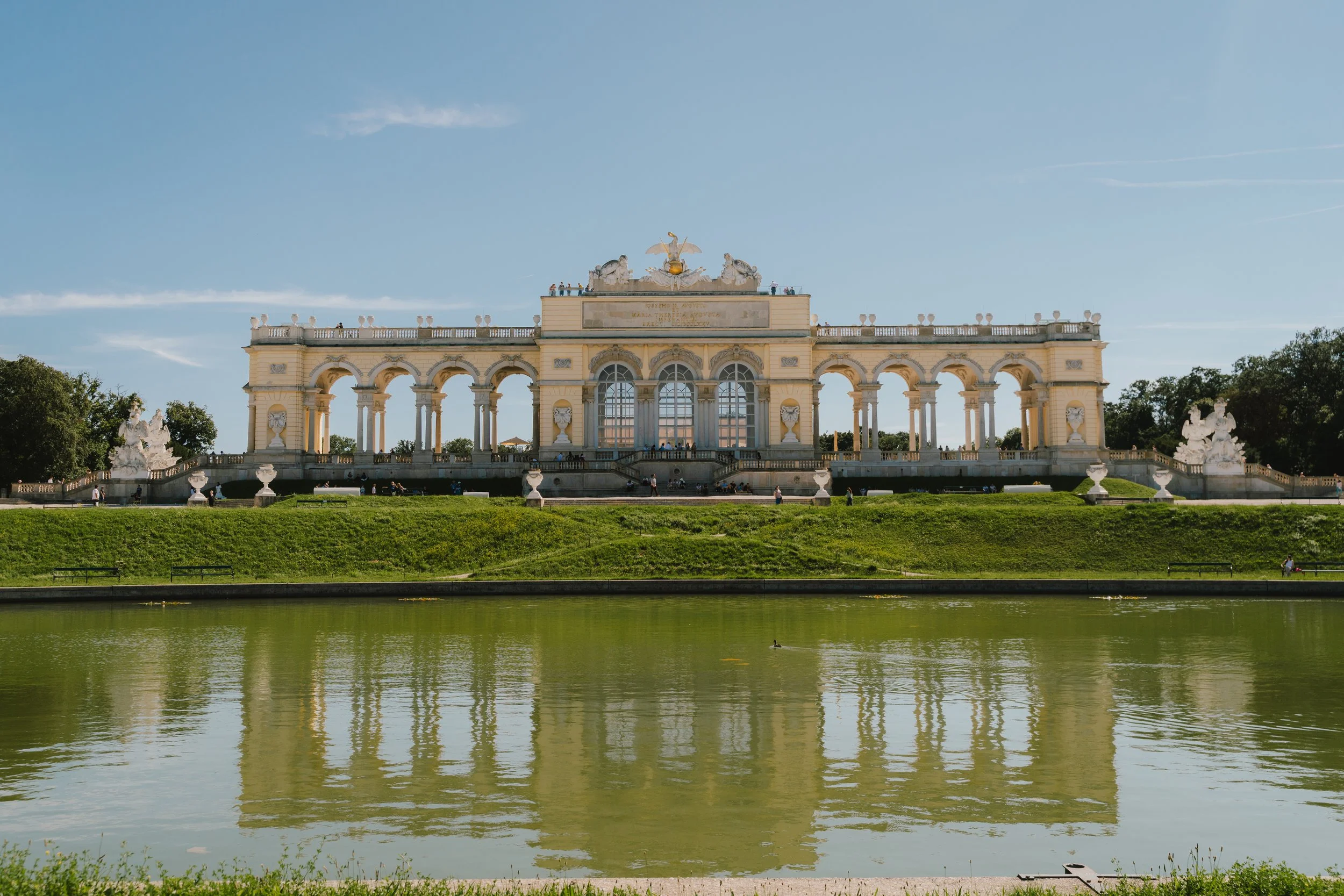 Serene park scene featuring an elaborate, cream-colored, neoclassical building with arches and sculptures, reflected in a calm body of water in the foreground, under a clear blue sky.