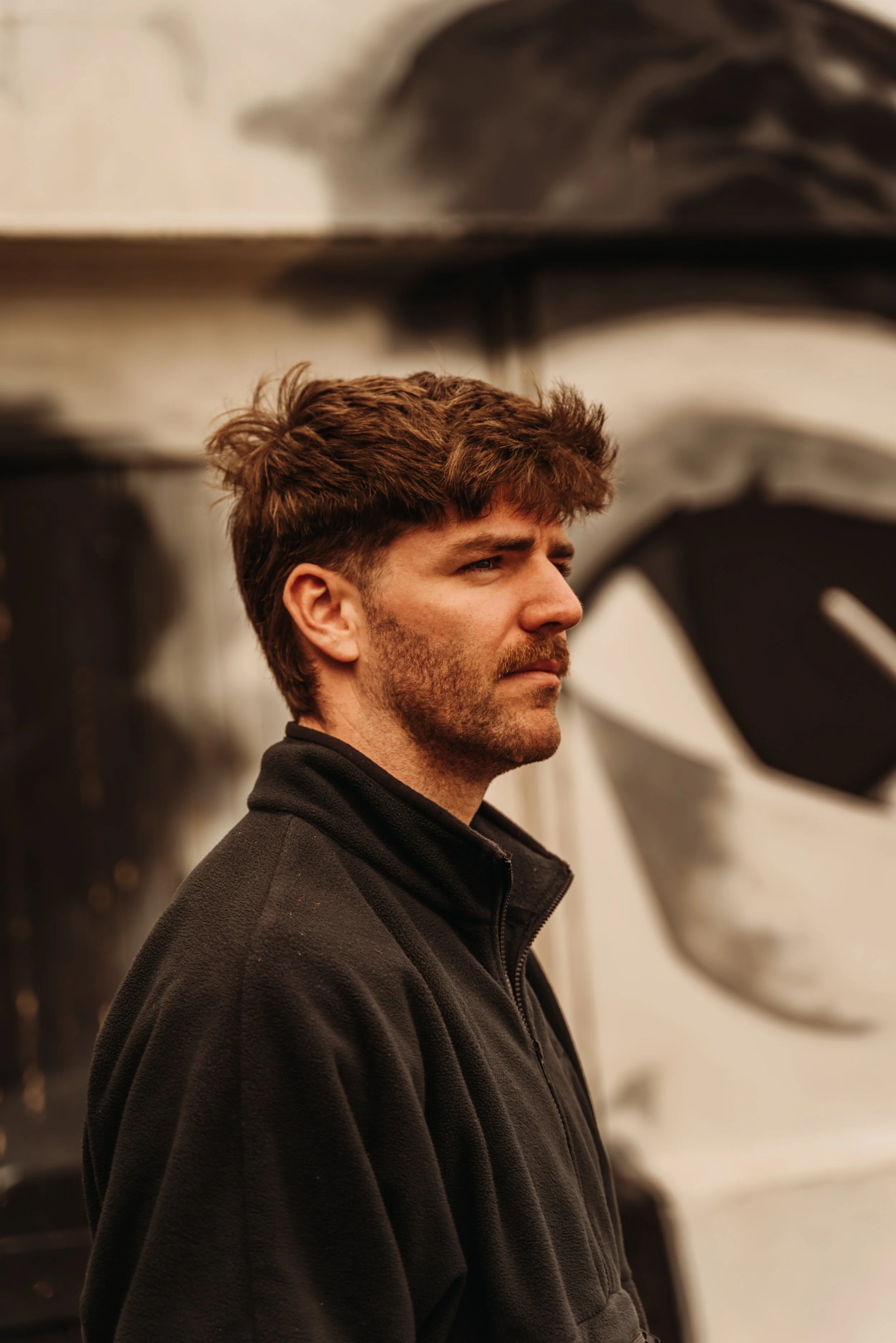 A man with brown hair and a beard, wearing a black fleece jacket, stands sideways and looks into the distance.