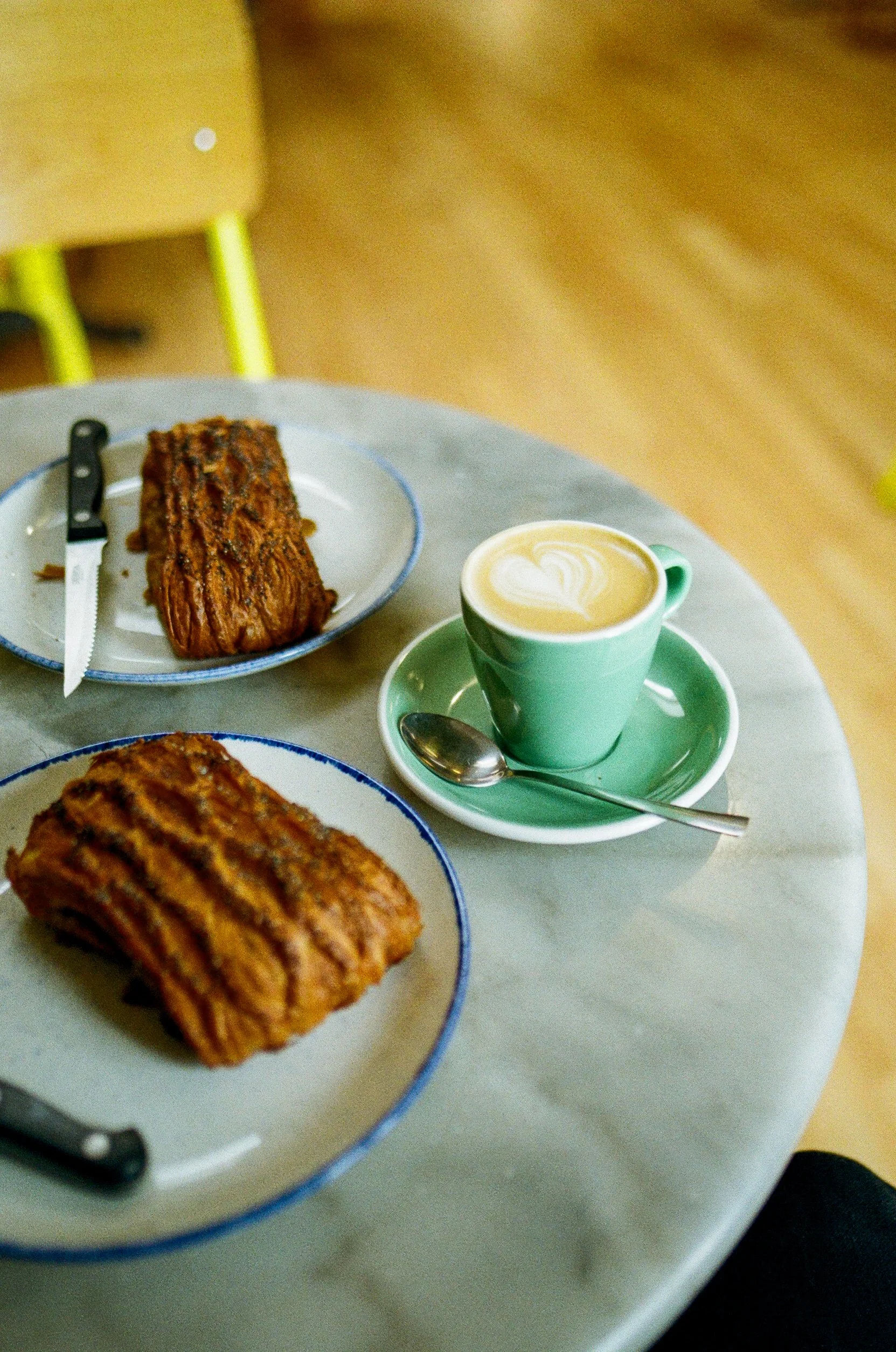 Two plates with baked goods, one with a piece of cake or pastry and the other with a roll, along with a green cup of latte with heart-shaped foam art and a spoon on a saucer, all on a white marble table.