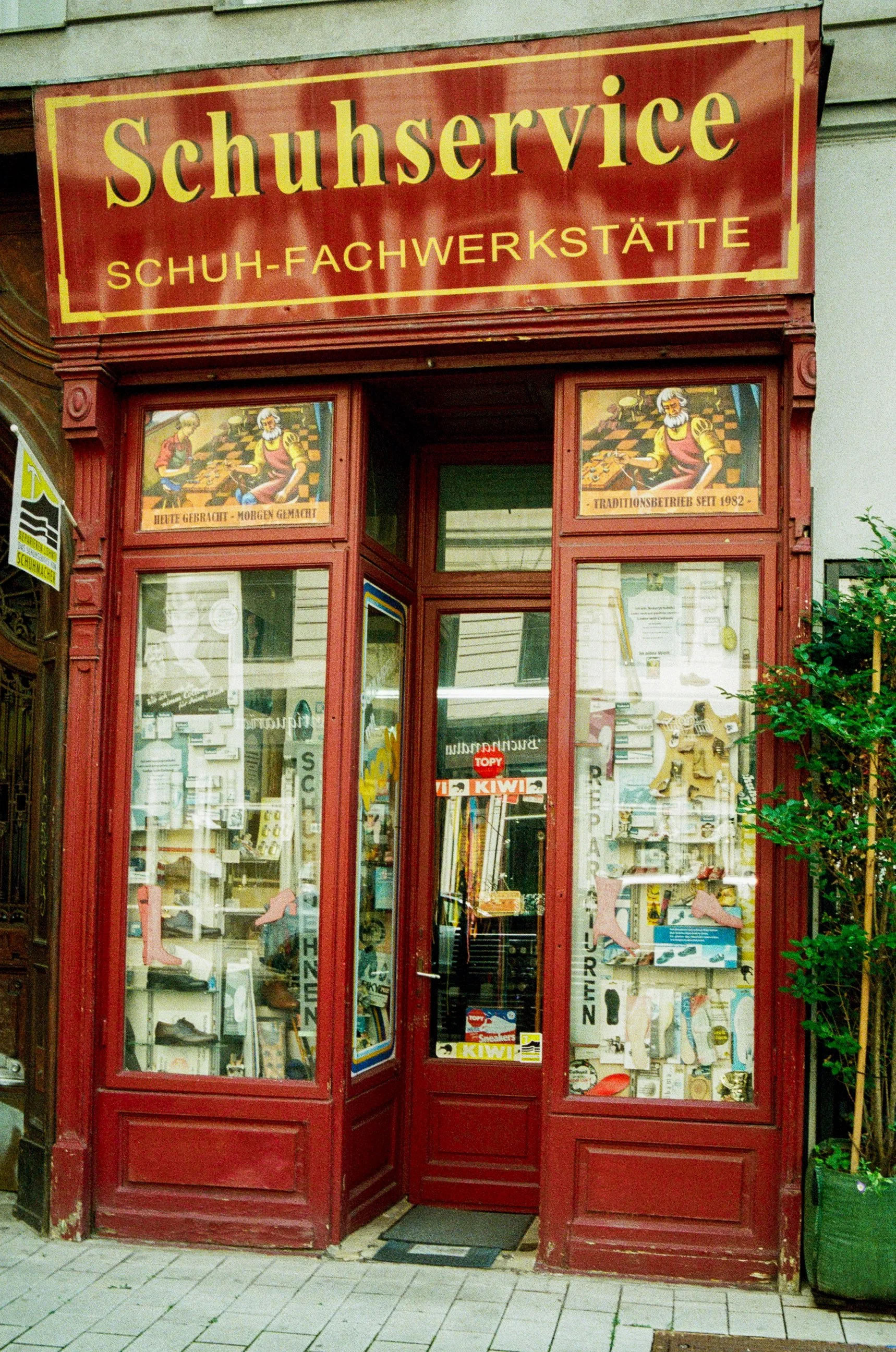 Red storefront for a shoe repair shop with signage in German, displaying shoes and shoe-related items in the window.