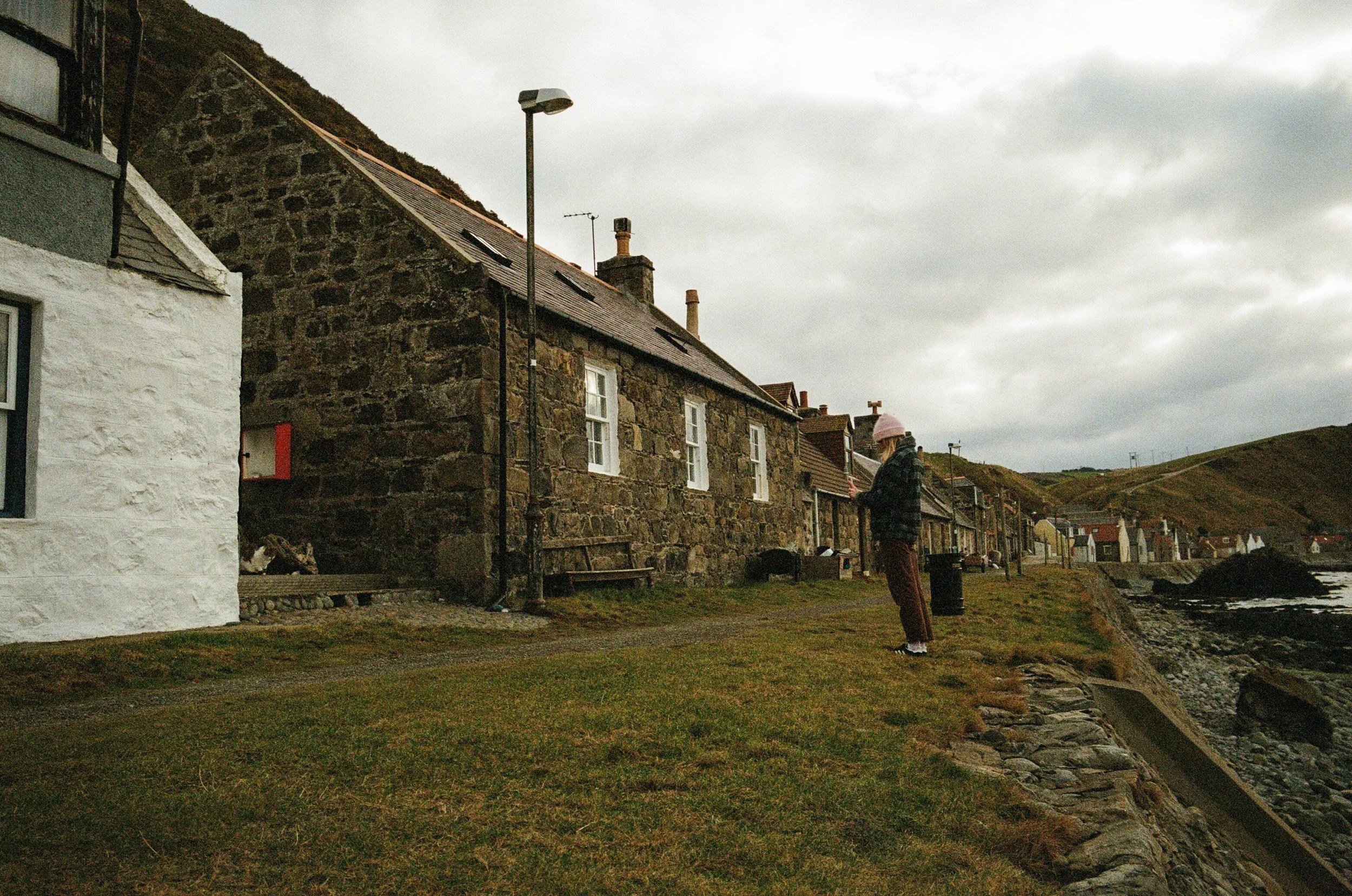 A person standing on the grass near the shoreline, looking at their phone, with stone and white-walled houses on a cloudy day.