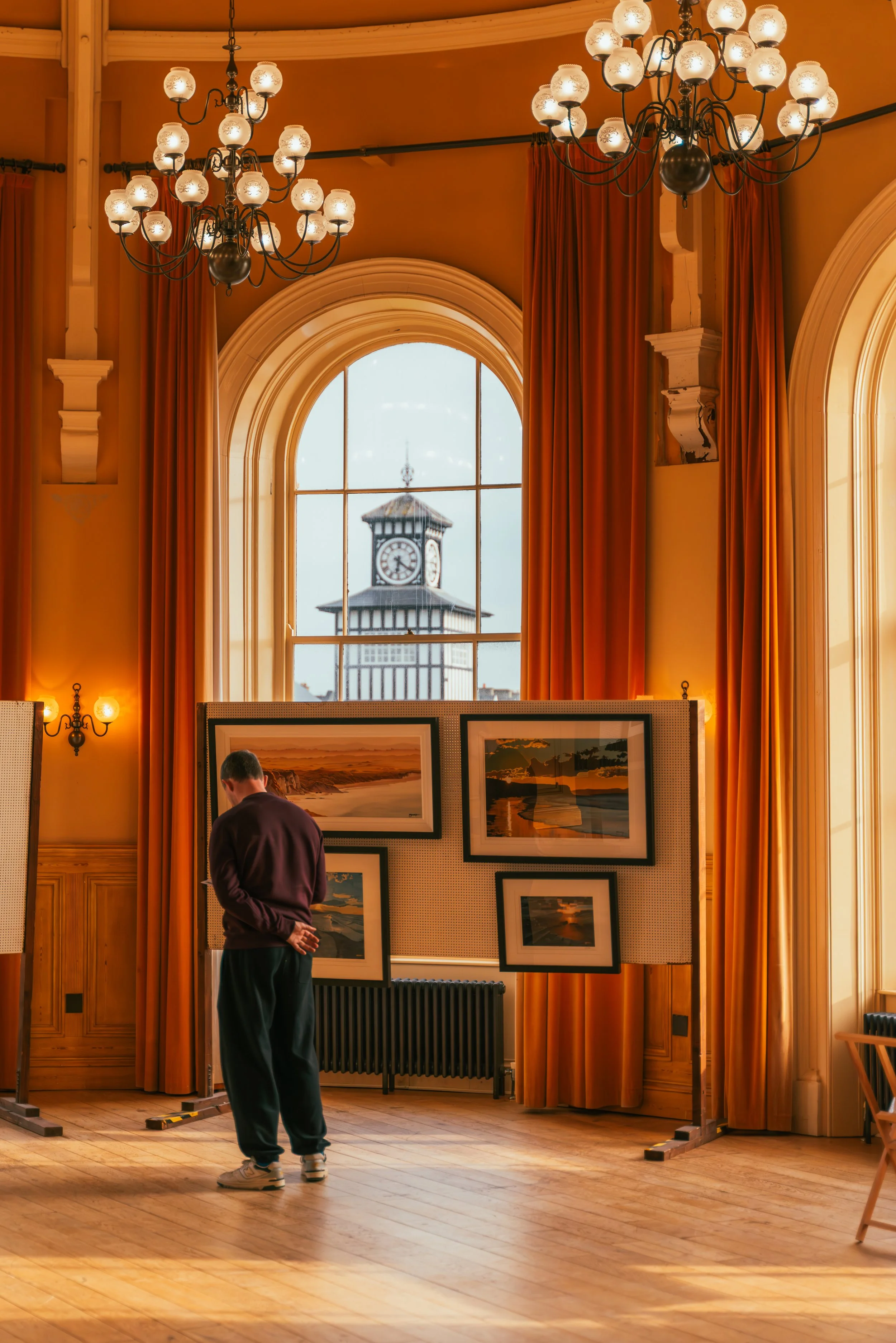 A person is viewing landscape paintings at an art exhibit inside a room with large arched windows, orange curtains, and chandeliers, with a clock tower visible outside through the windows.