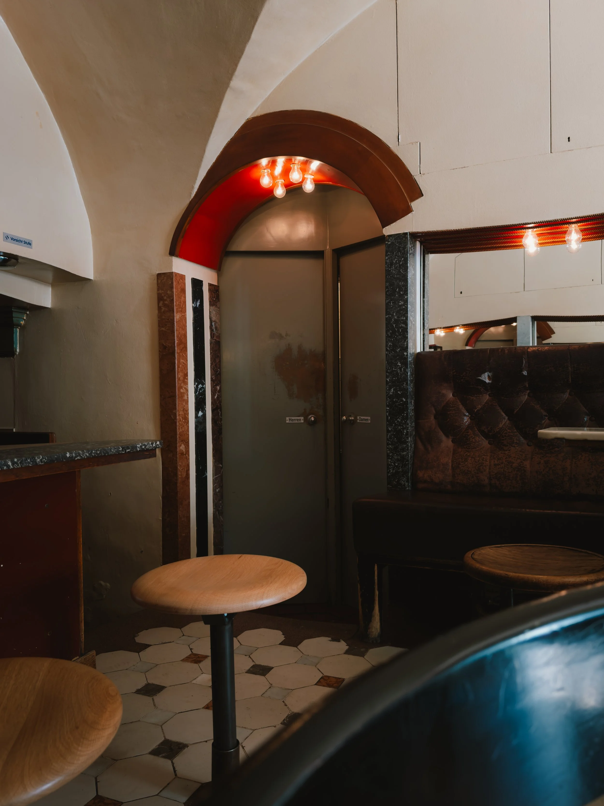 Interior of a café or restaurant with wooden stools, leather benches, hexagonal tiled floor, and a metallic door with signs, illuminated by small light bulbs.