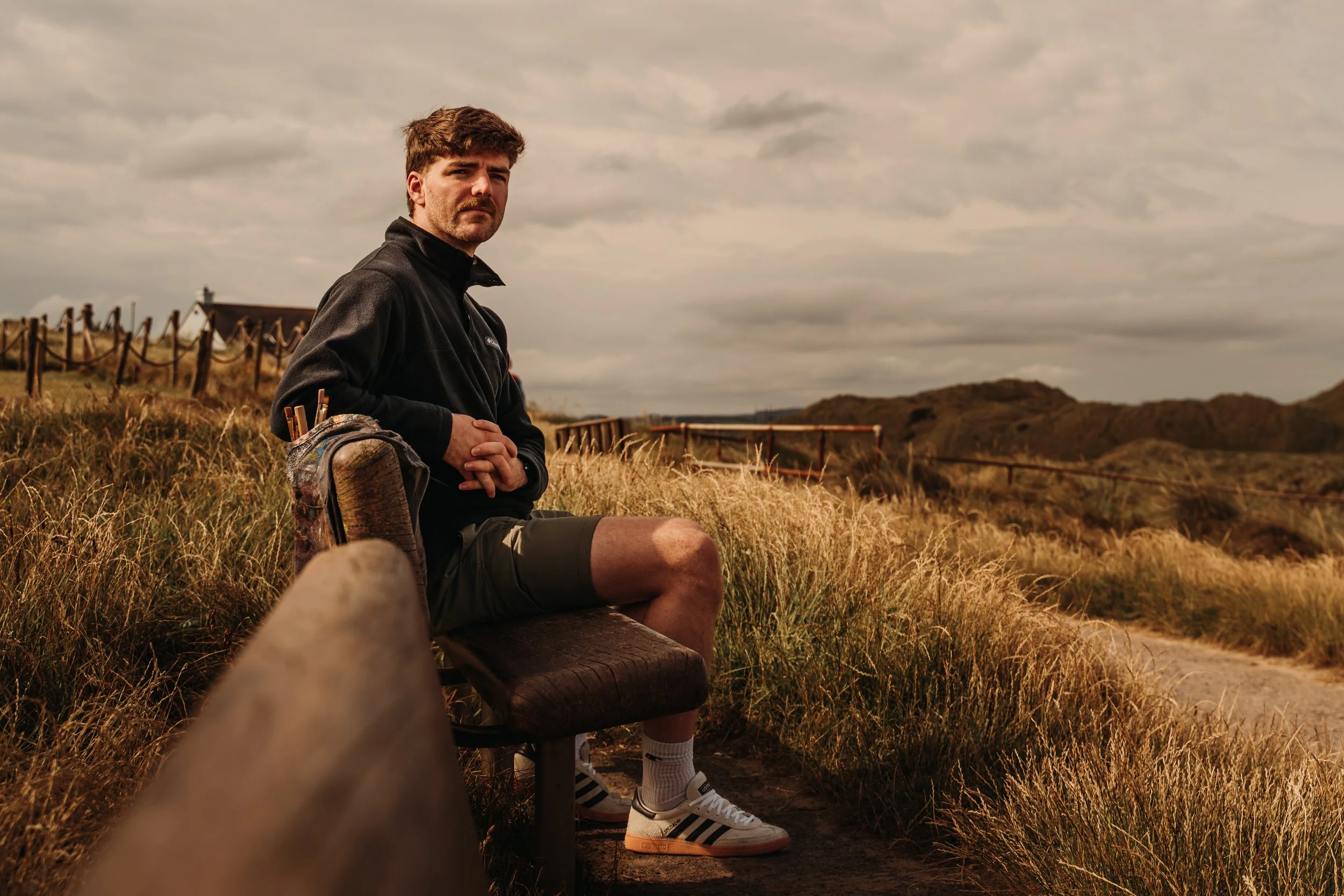 A man sitting on a bench outdoors in a grassy area on a cloudy day, with hills and a wooden fence in the background.