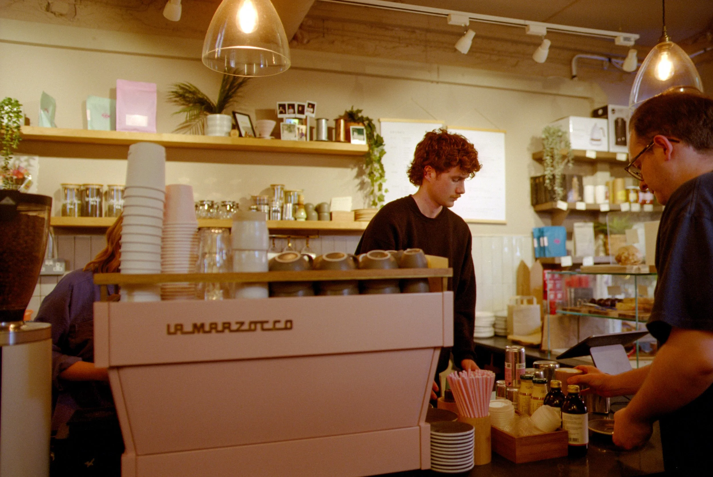 A barista with curly red hair and a black shirt stands behind the counter of a coffee shop, preparing an order. The shop has warm lighting, wooden shelves with various jars and packages, a coffee machine labeled 'La Marzocco,' cups, and a customer wi