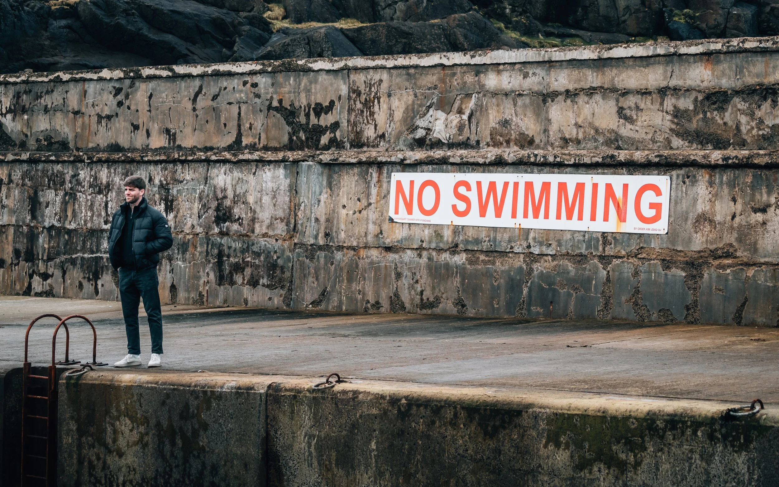 A man in a black jacket and white sneakers standing on a concrete dock with a rusty ladder, next to an aged stone wall with a large 'No Swimming' sign.