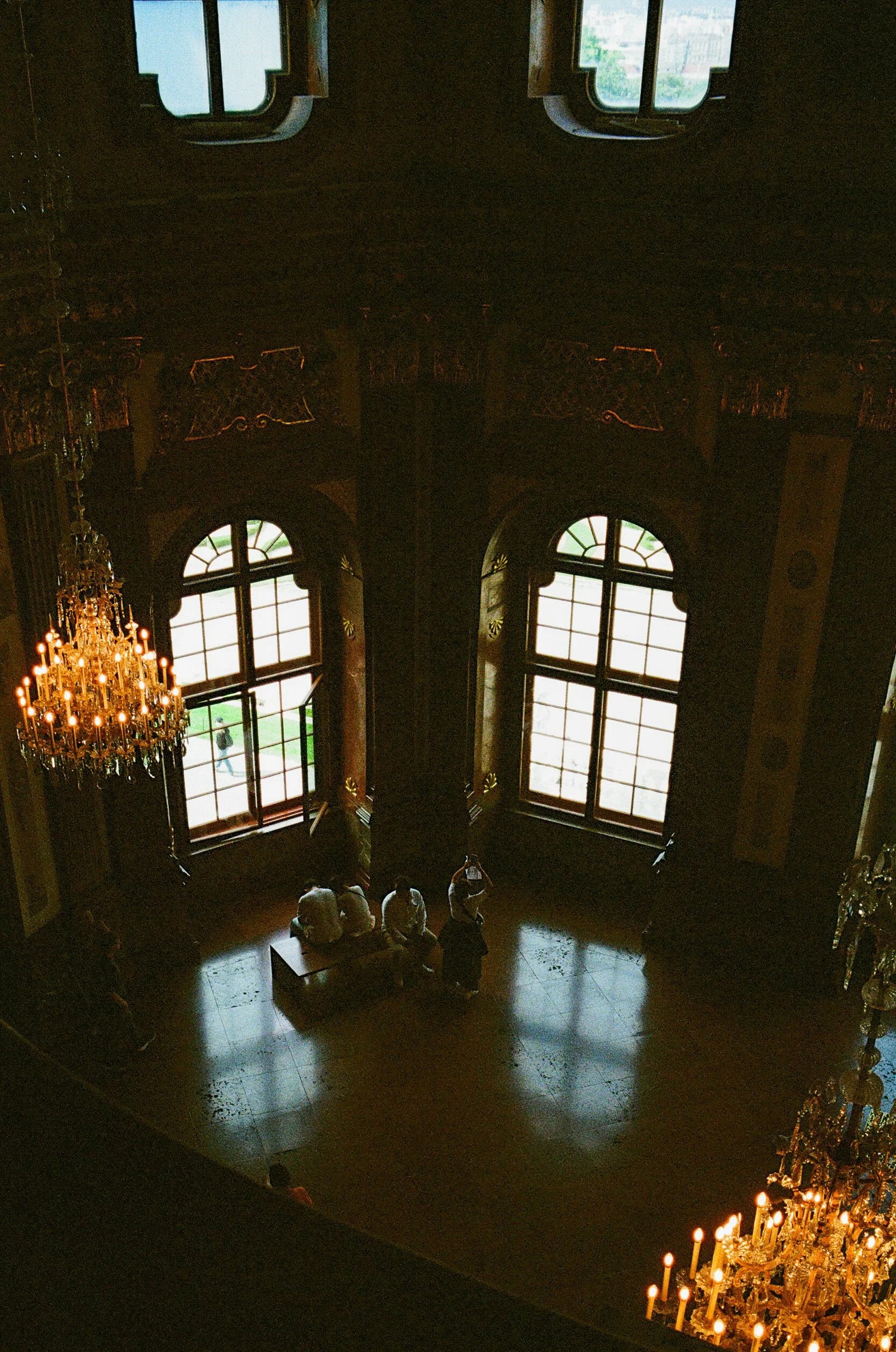 Interior of a large, ornate room with tall arched windows, chandeliers, and decorative woodwork. A group of people are sitting and taking photos near the windows.