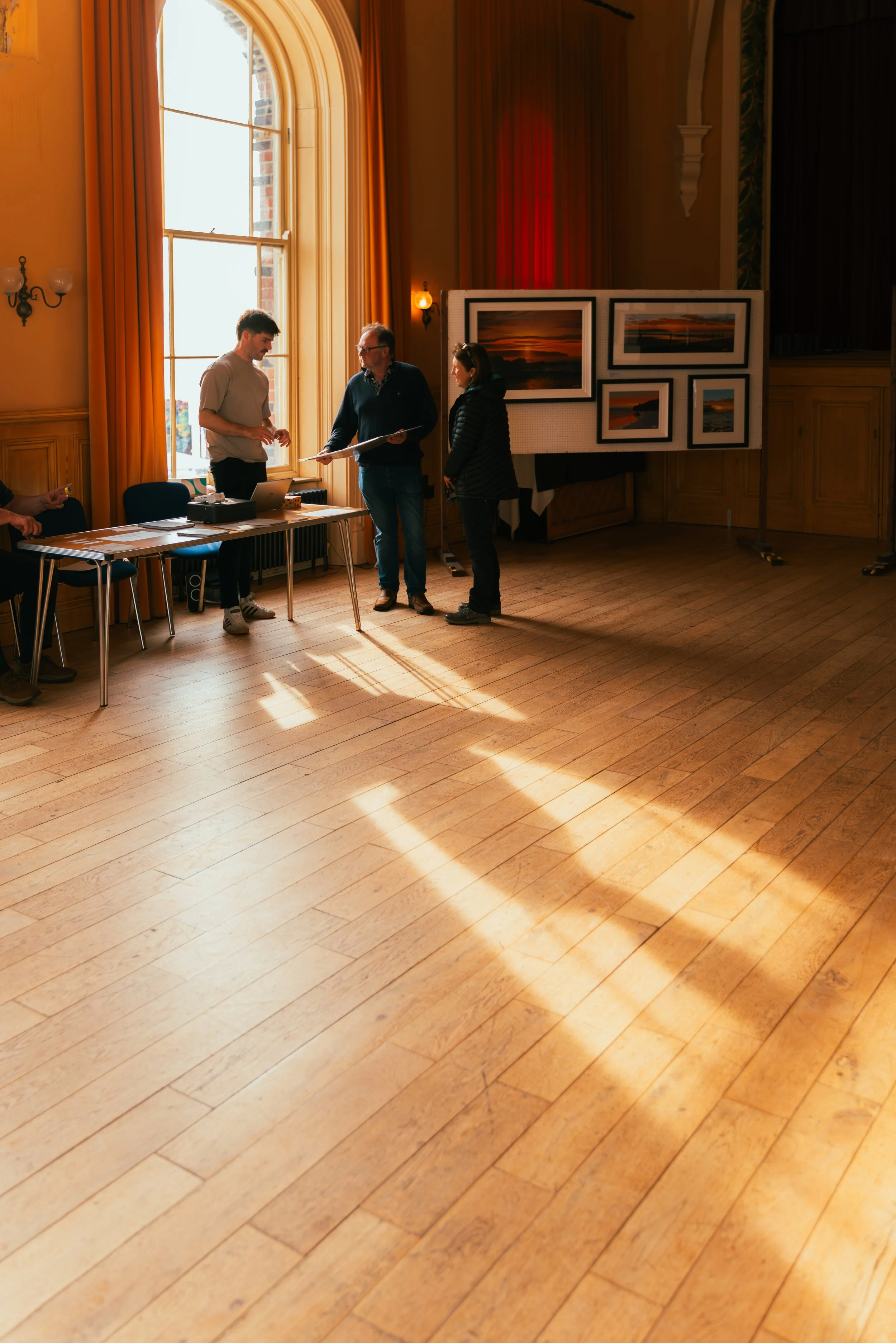 Three people standing and talking near a table and an art display in a sunlit room with a large window and wooden floors.