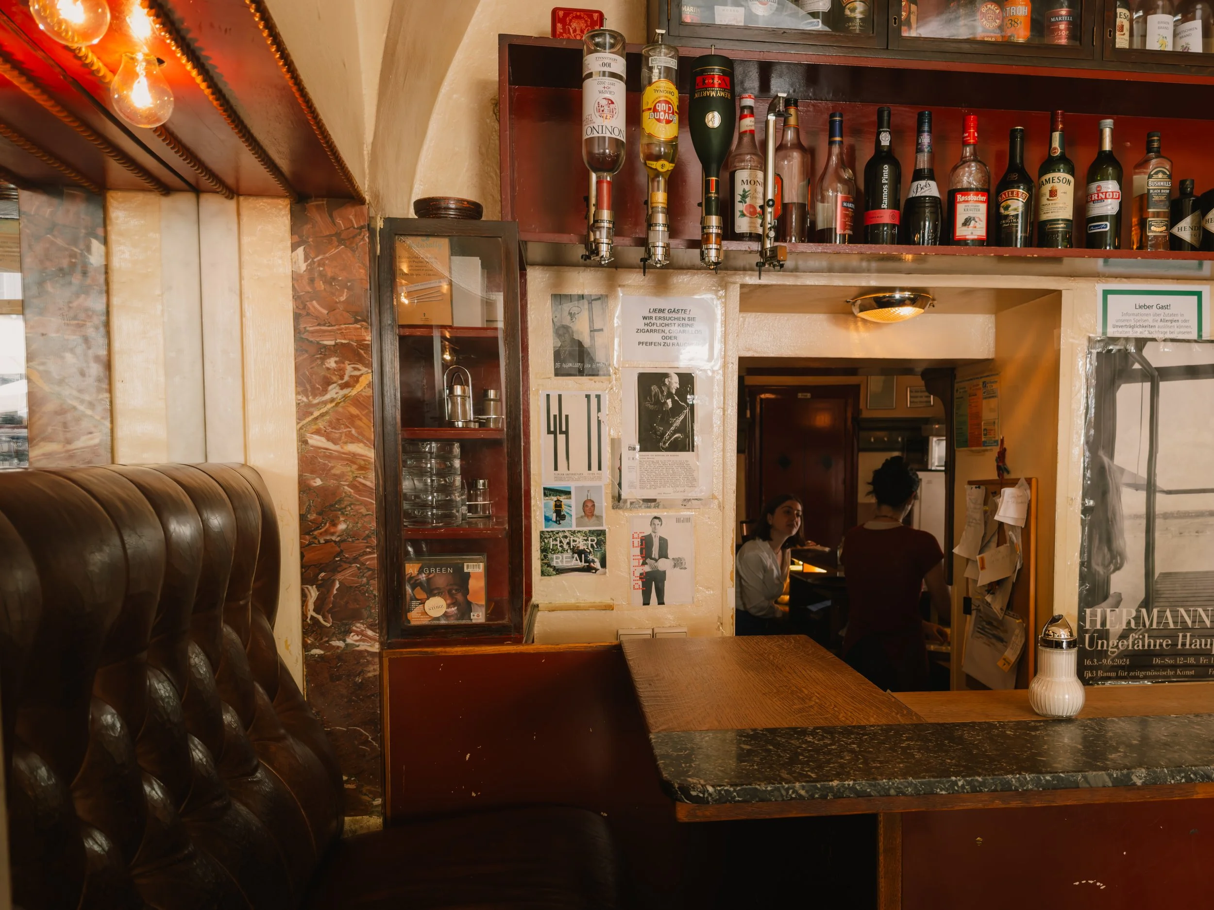 Interior of a cozy bar or cafe with leather seating, wooden and marble counters, and shelves of bottles and glasses. Two women are sitting at a table in the background, engaging in conversation.