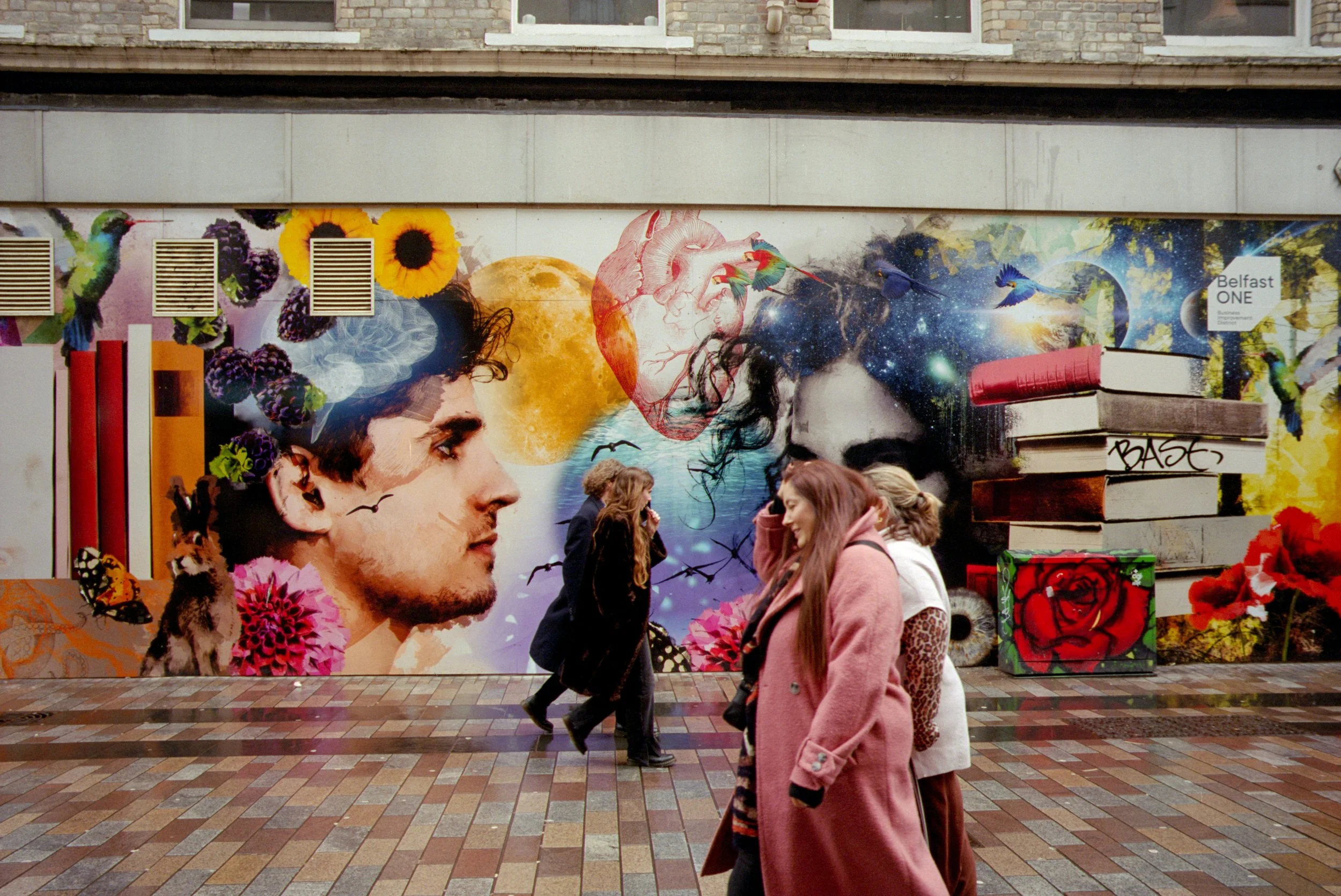 Street scene with a colorful mural featuring a man's profile, a woman's profile, books, sunflowers, an illustration of a heart, space elements, and various animals. Several pedestrians walk in front of the mural on a brick sidewalk.