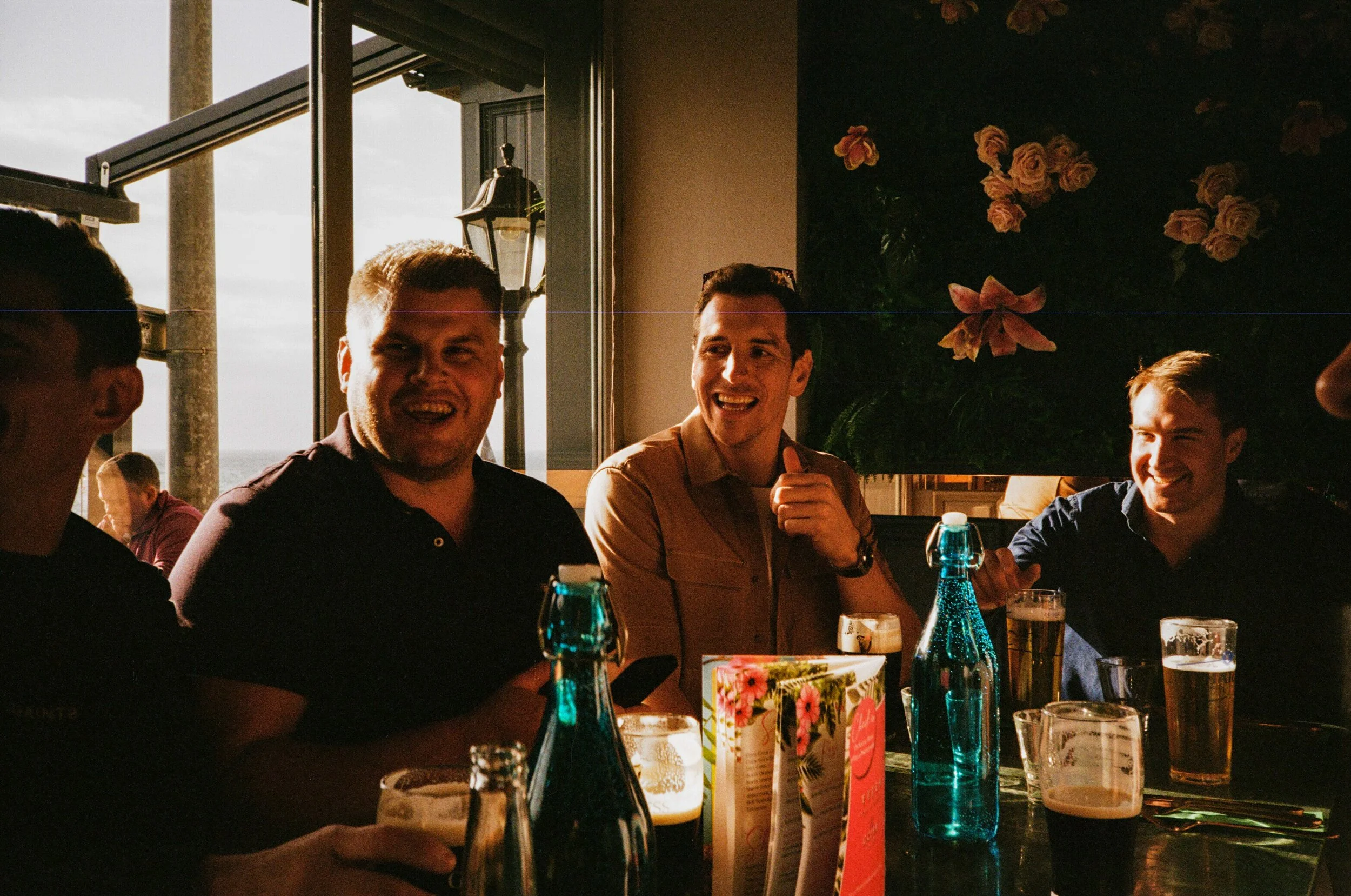 A group of young men enjoying drinks and conversation at a restaurant or bar with large windows and a floral wall decoration in the background.