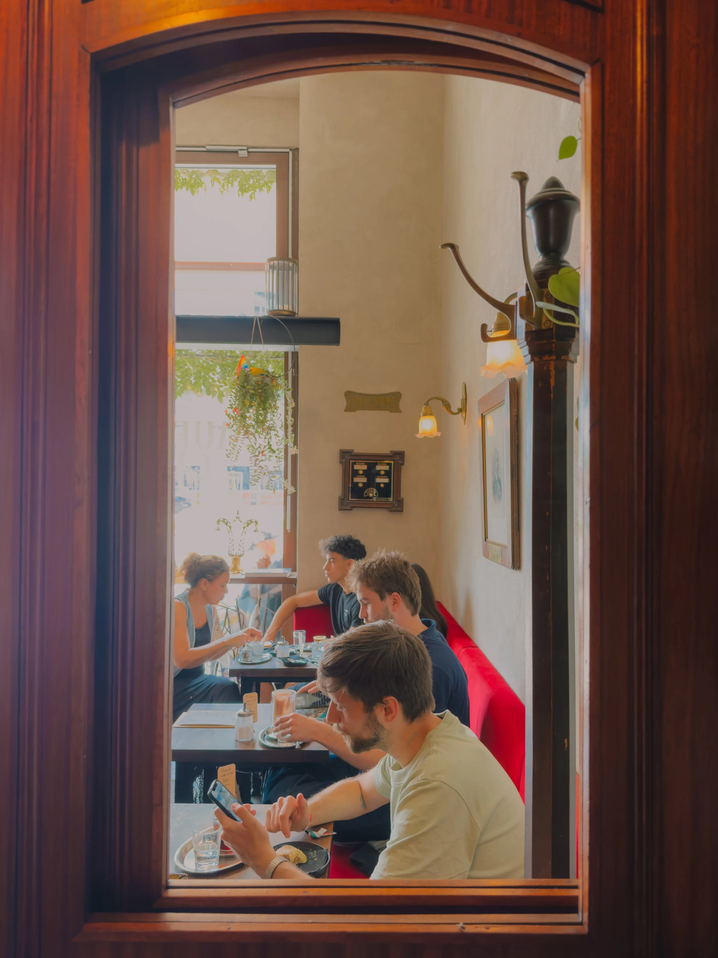 View through a wooden window frame into a cozy restaurant with a group of people dining, some using their phones, with natural light coming from large windows.