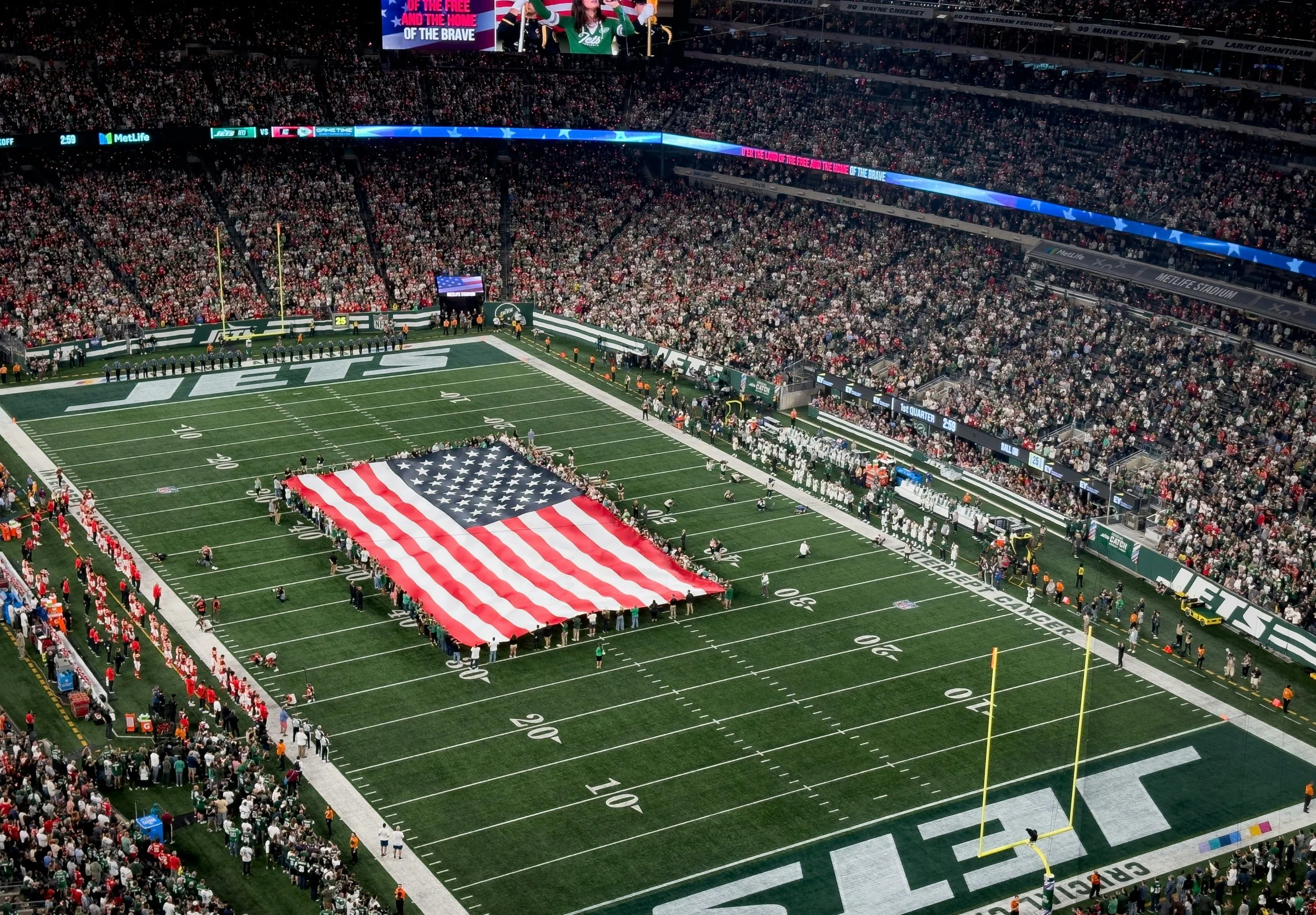 A large American football stadium filled with spectators, with a giant American flag laid on the field, and players and officials standing around it before a game. The field has markings for yard lines and is decorated with team logos and advertisements.