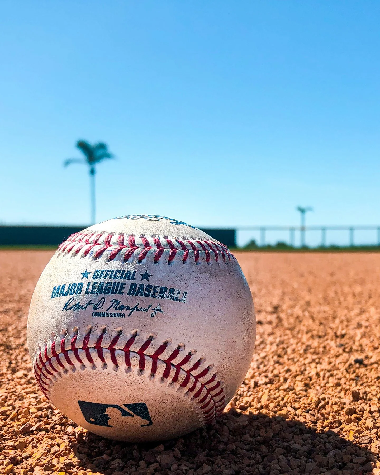 Close-up of a baseball on a dirt field with a clear blue sky, two light poles, and a fence in the background.