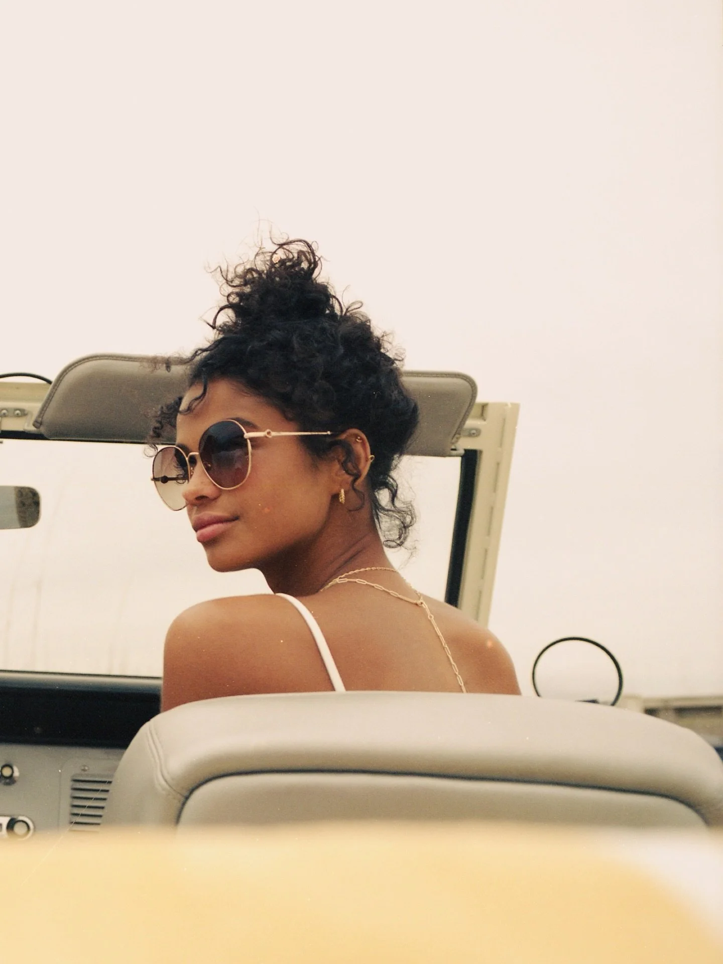 A woman wearing round sunglasses, gold earrings, and a thin gold necklace, sitting in front of a mirror with her head slightly turned to the side, in what appears to be a salon or bathroom setting.