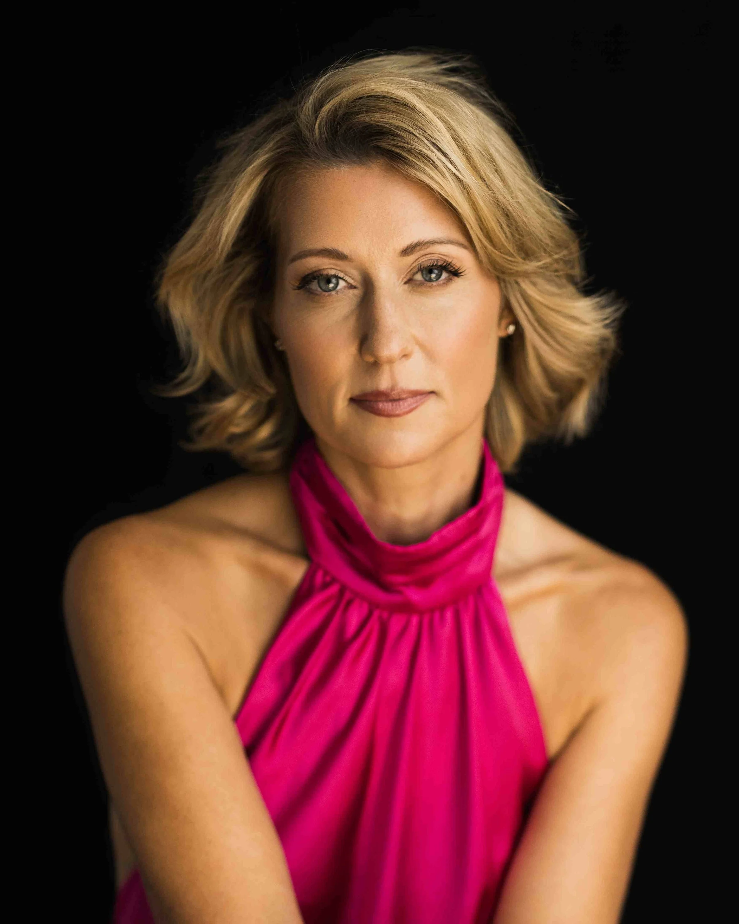A woman with shoulder-length blonde hair wearing a pink halter dress, posing against a black background.