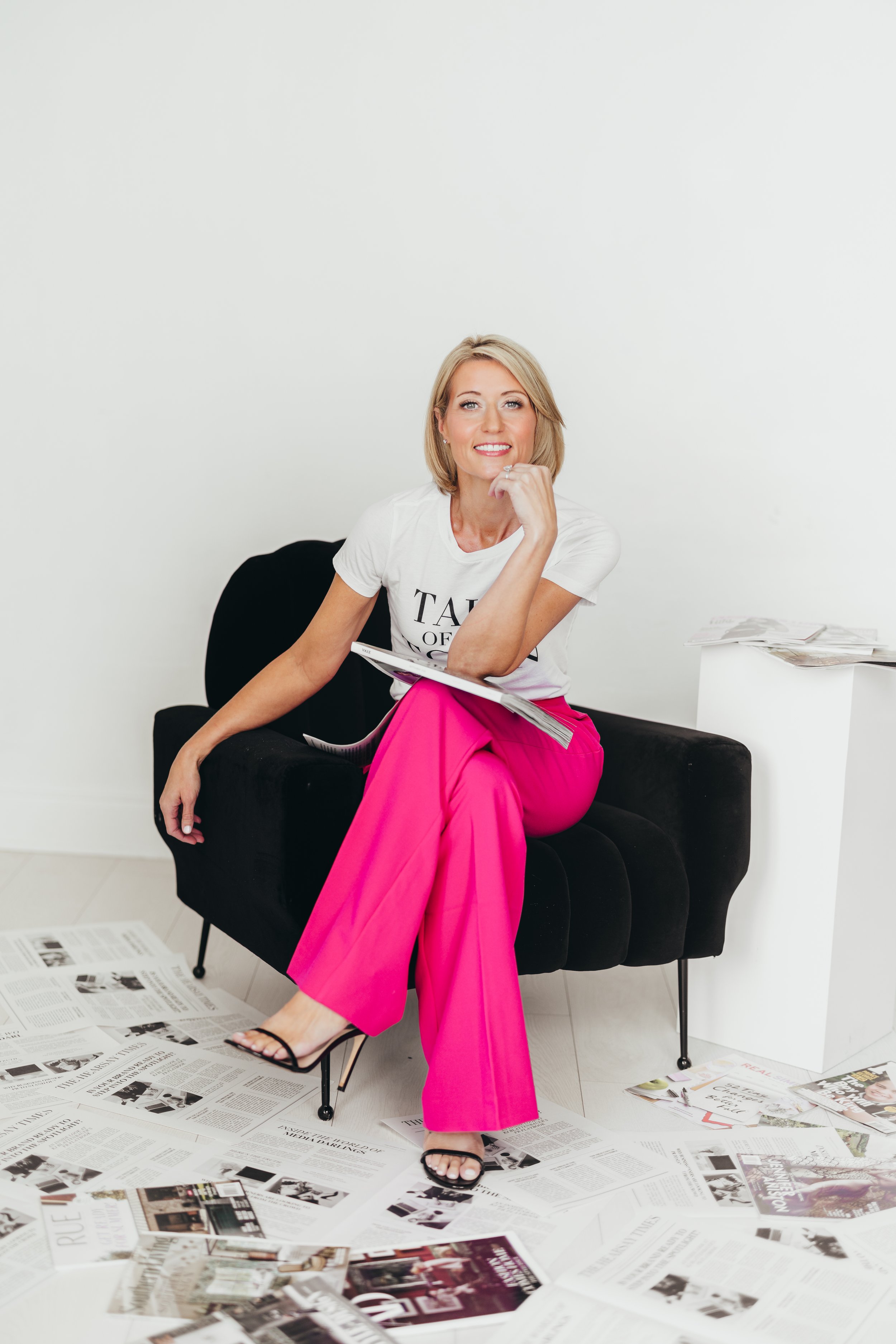 A woman with blonde hair wearing a white t-shirt and bright pink pants sits on a black chair with newspapers scattered on the floor and on a small white table beside her. She is smiling and resting her chin on her right hand.