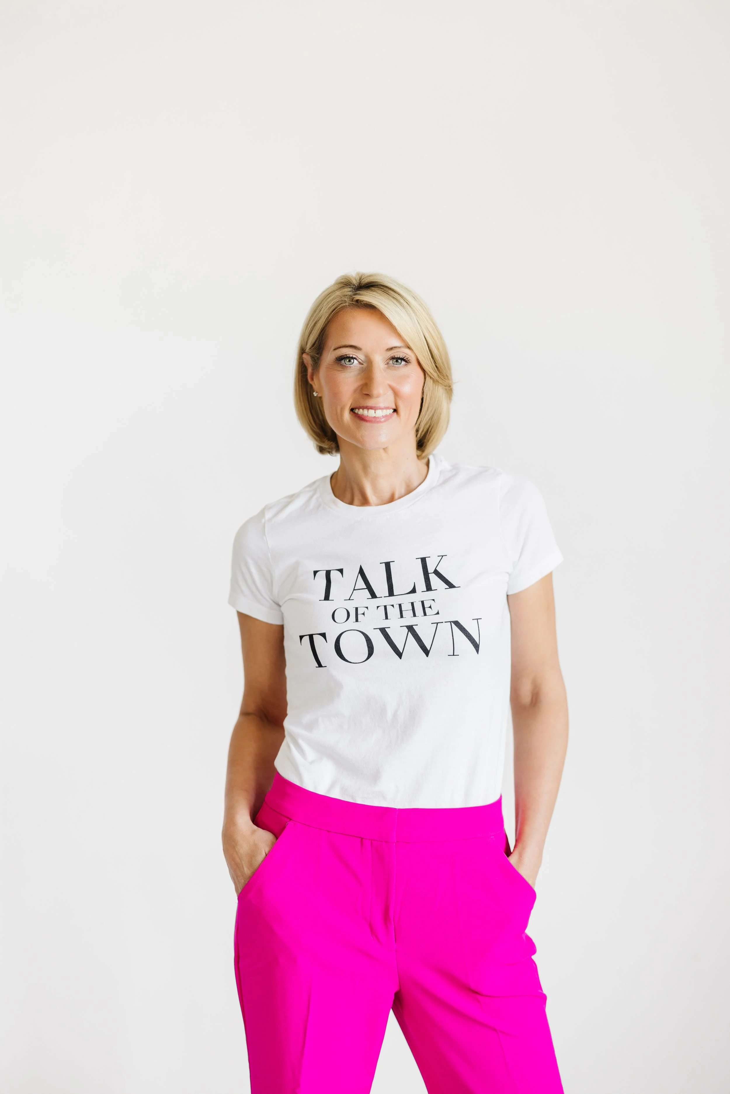 A woman with blonde hair wearing a white T-shirt that says 'TALK OF THE TOWN' and bright pink pants, standing against a plain white background.