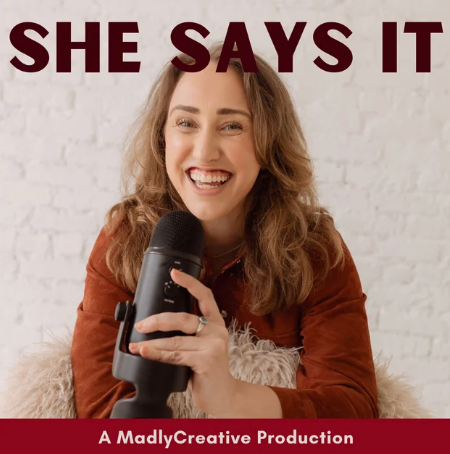 A young woman with long curly hair smiling and holding a microphone in front of a white brick wall. Text above her says 'SHE SAYS IT' and below says 'A MadlyCreative Production'.