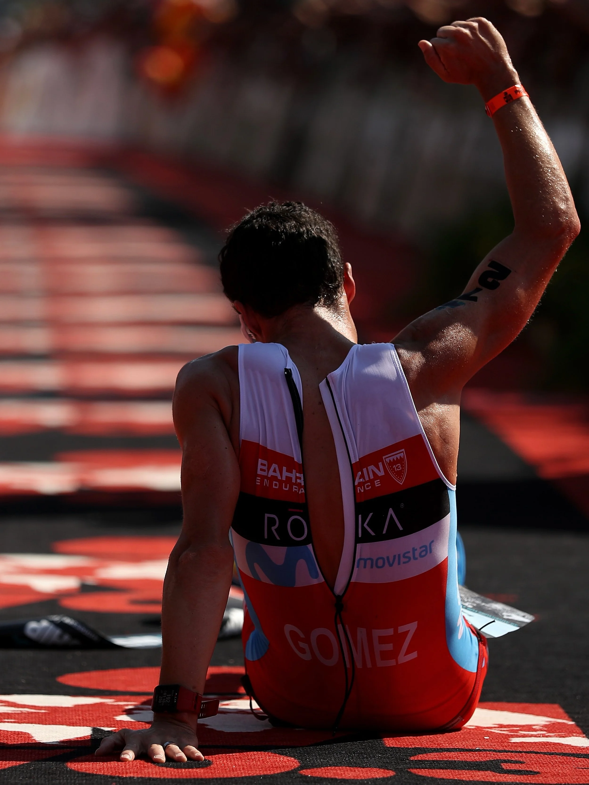 Athlete sitting on the ground after a race, wearing a red, white, and blue uniform with the name Gomez on the back, raising one arm. The athlete is sweaty and appears exhausted.