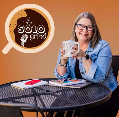 A smiling woman with glasses holding a coffee mug sitting at a black table with notebooks and a red phone, with a logo for 'the solo grind' podcast in the background.